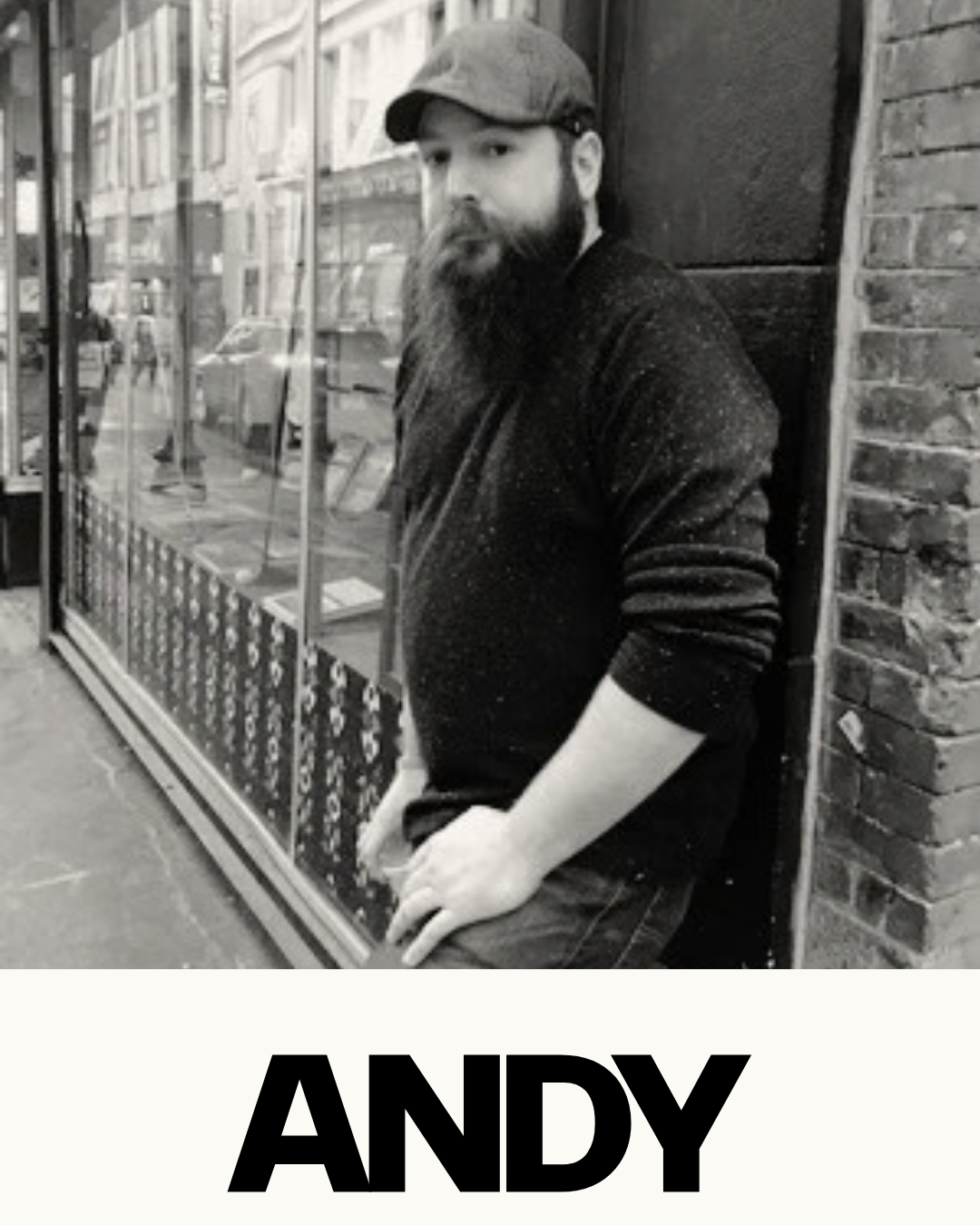 A black and white photo of a man with a beard wearing a flat cap and dark sweater leaning against a brick wall outside a storefront with glass windows, with the name "Andy" displayed at the bottom.