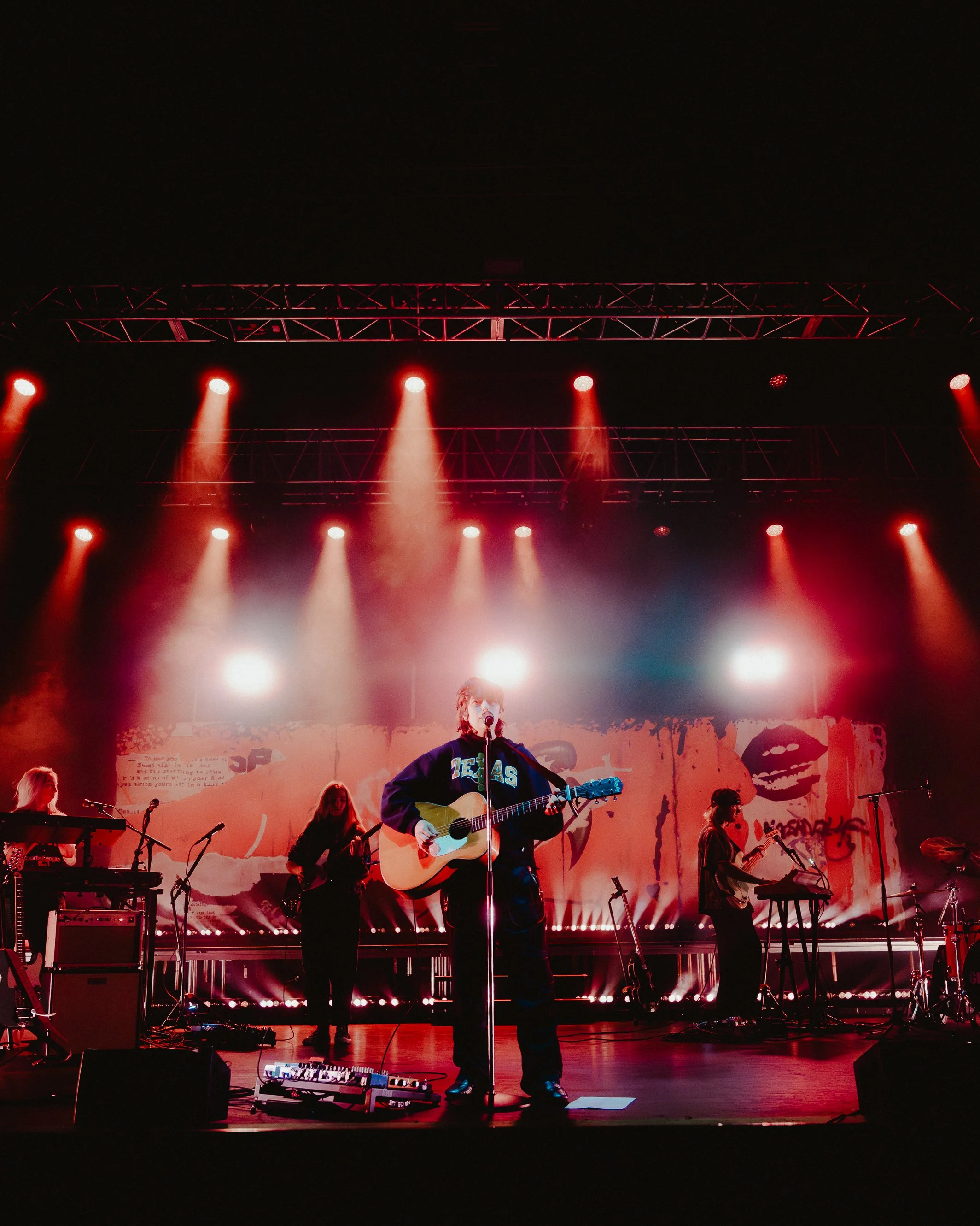 Band performing on stage with red lighting, including a guitarist singing into a microphone, keyboard players, and other musicians, with a backdrop featuring graphics and text.