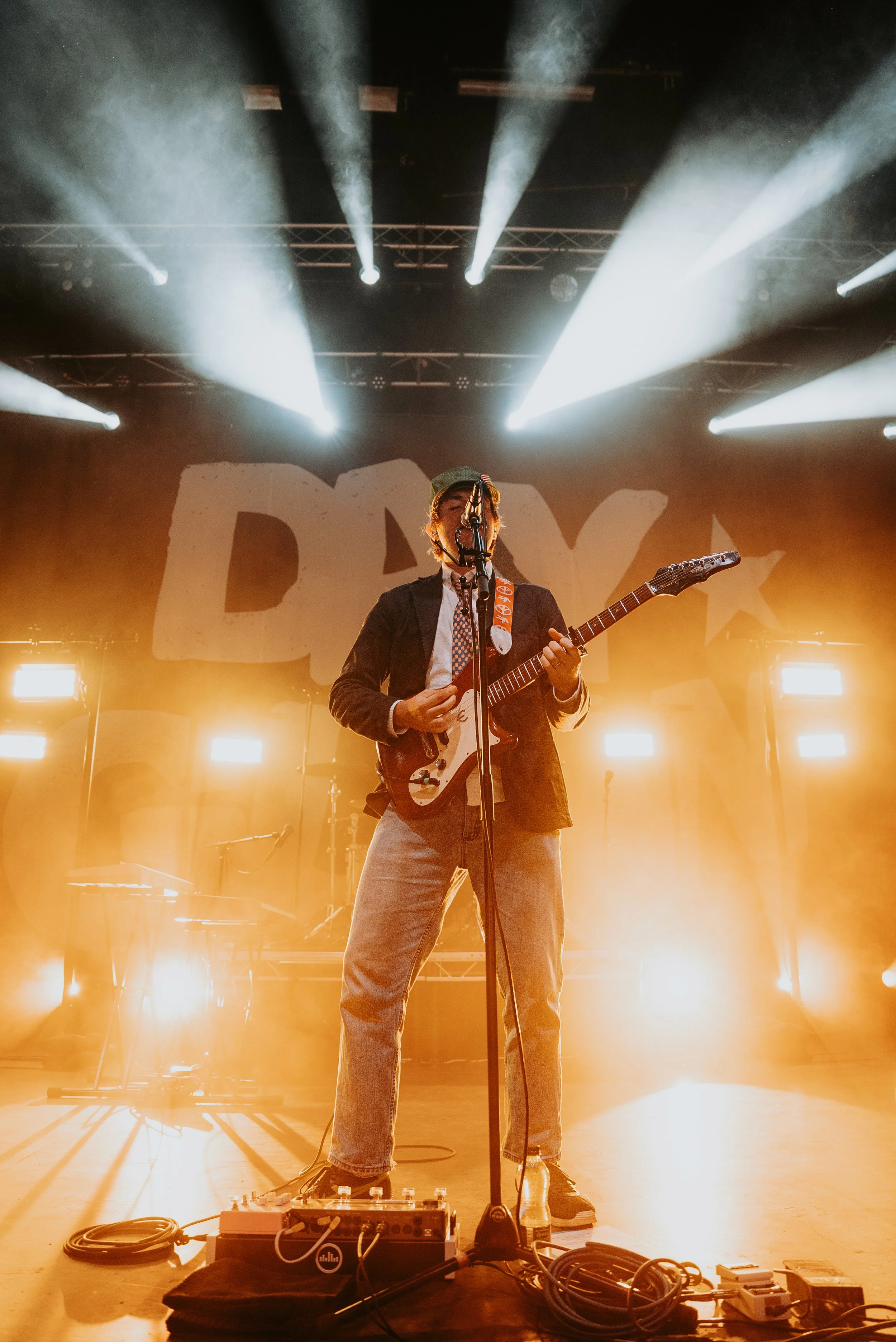 A musician playing an electric guitar on stage with bright stage lights and a large backdrop with partially visible letters.
