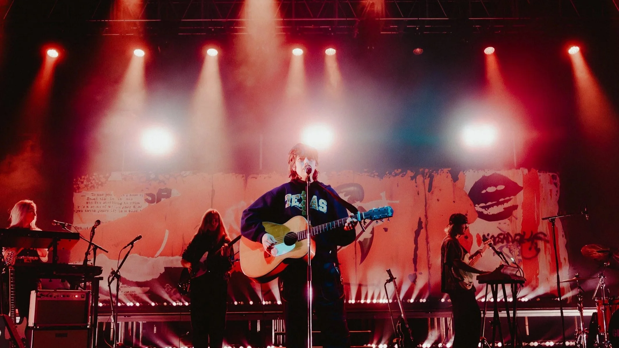A band performing on stage with a singer playing guitar and singing into a microphone. The stage is lit with red and white lights, and there is a backdrop with graffiti art and large lips.