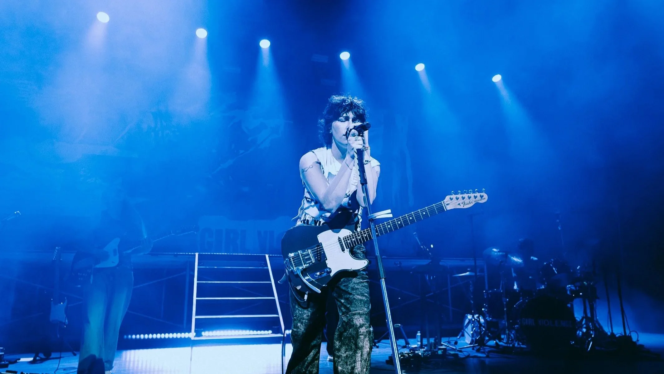 Woman with short curly hair singing into a microphone while playing an electric guitar on stage, illuminated by blue stage lights.