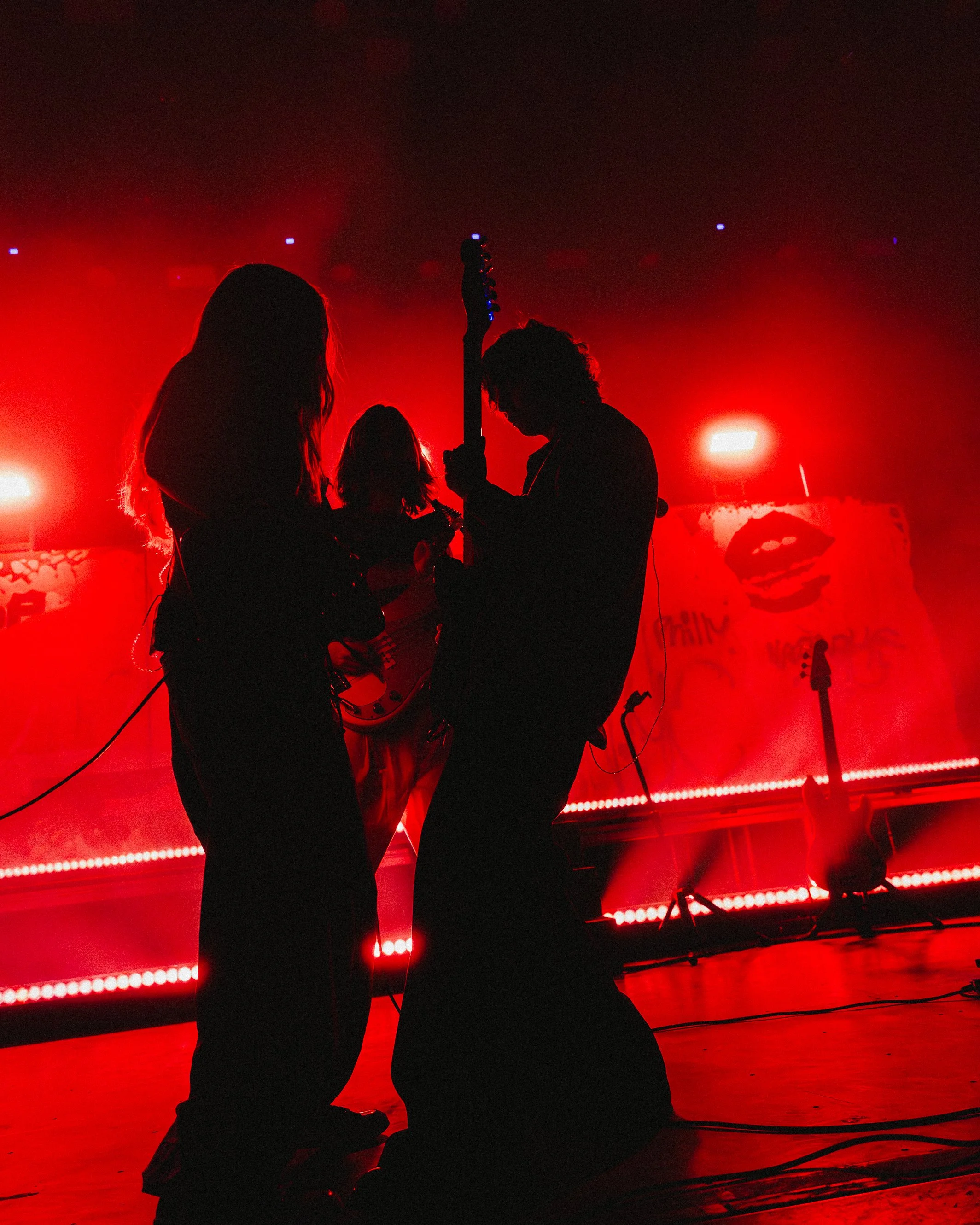 Silhouettes of four musicians on stage with red lighting, one playing a guitar and another with a microphone, in front of a backdrop with graphics and text.