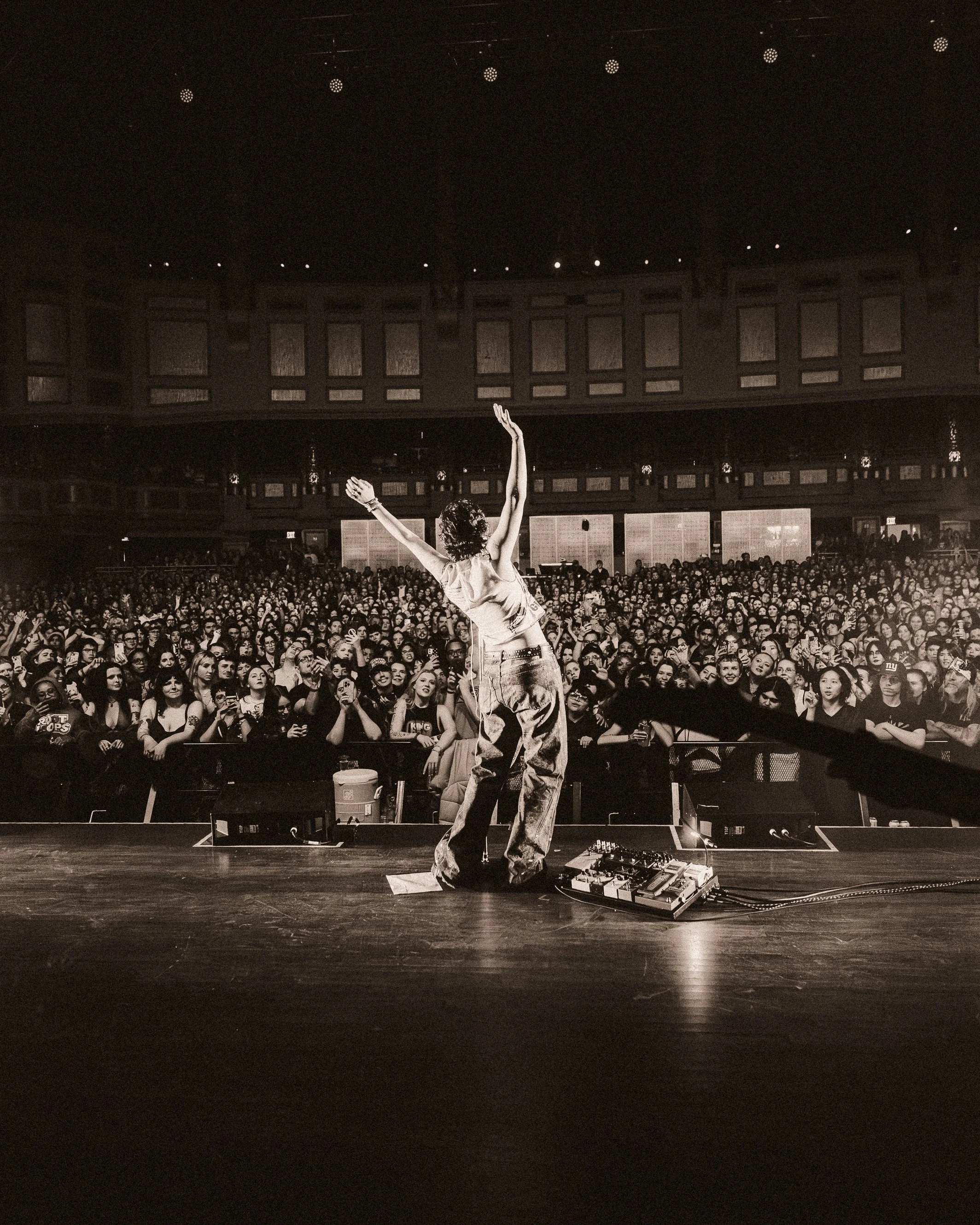 A performer on stage at a concert, facing a large audience, with their arms raised. The venue has tiered balconies and stage equipment visible in the foreground, in black and white.