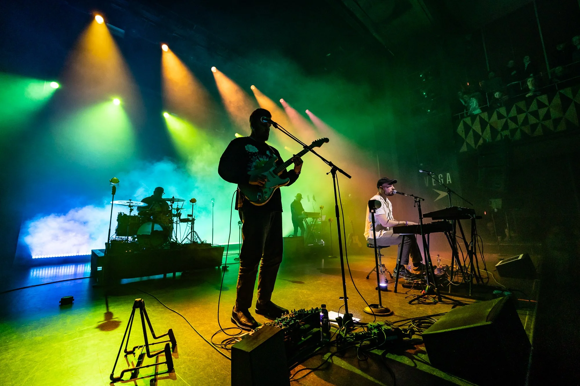 Musicians performing on stage with colorful lighting, including a guitarist, drummer, and keyboard player.