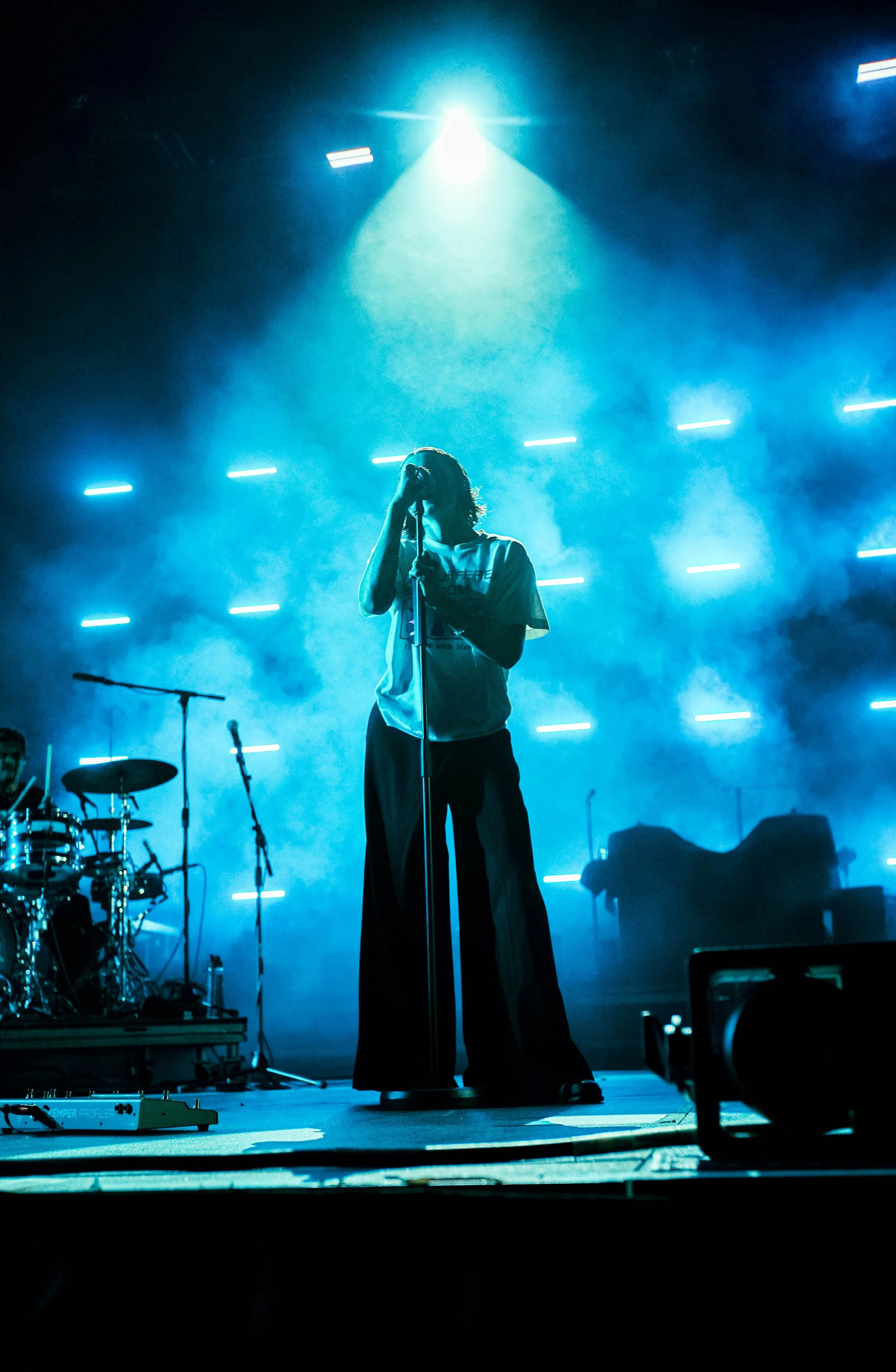 A person singing on stage with blue lighting, surrounded by musical instruments and equipment.