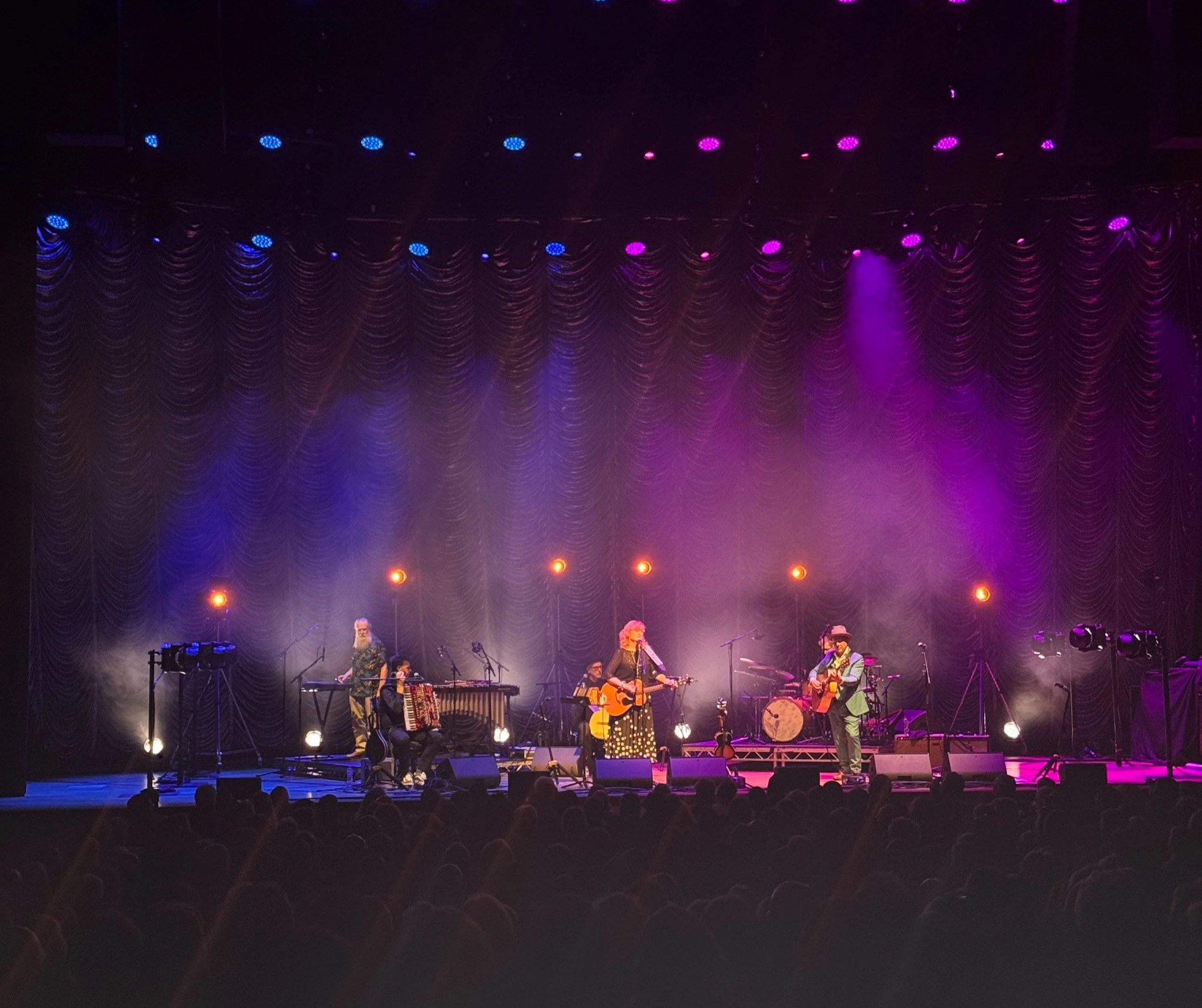 A musical concert stage illuminated with purple and blue lights. Several musicians are performing, including a woman in a black dress with white polka dots singing into a microphone, playing guitar, and other musicians playing instruments like drums, and keyboards. The audience is visible at the bottom.