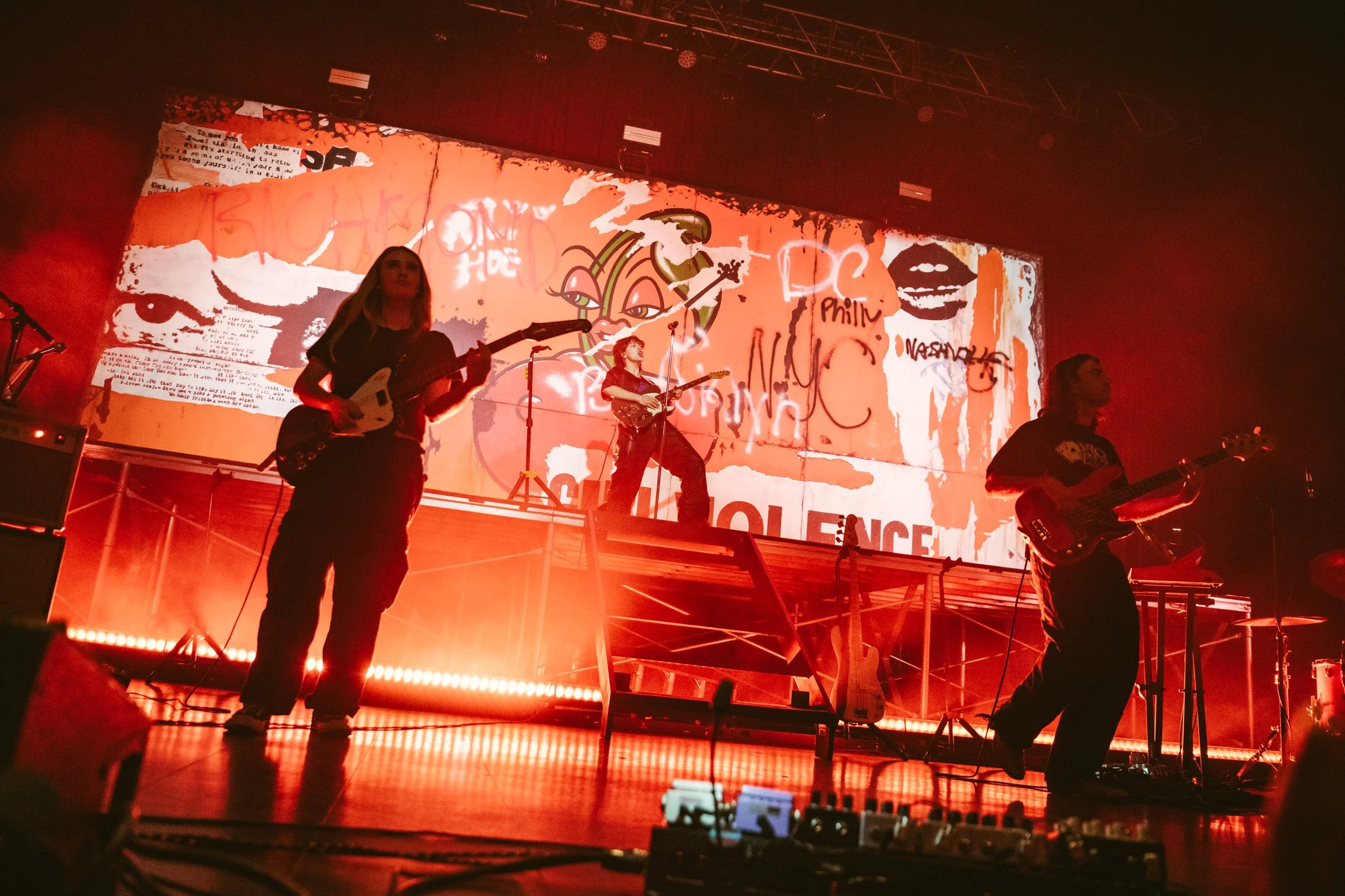 A band performs live on stage with guitars and microphones, illuminated by reddish stage lighting, in front of a large screen displaying graffiti-style artwork and text.