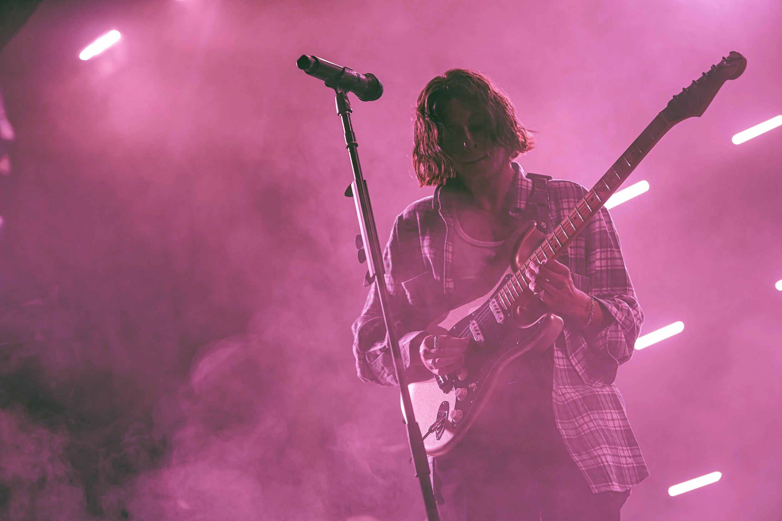 A person playing an electric guitar on stage with pink lighting and a stand with a microphone.