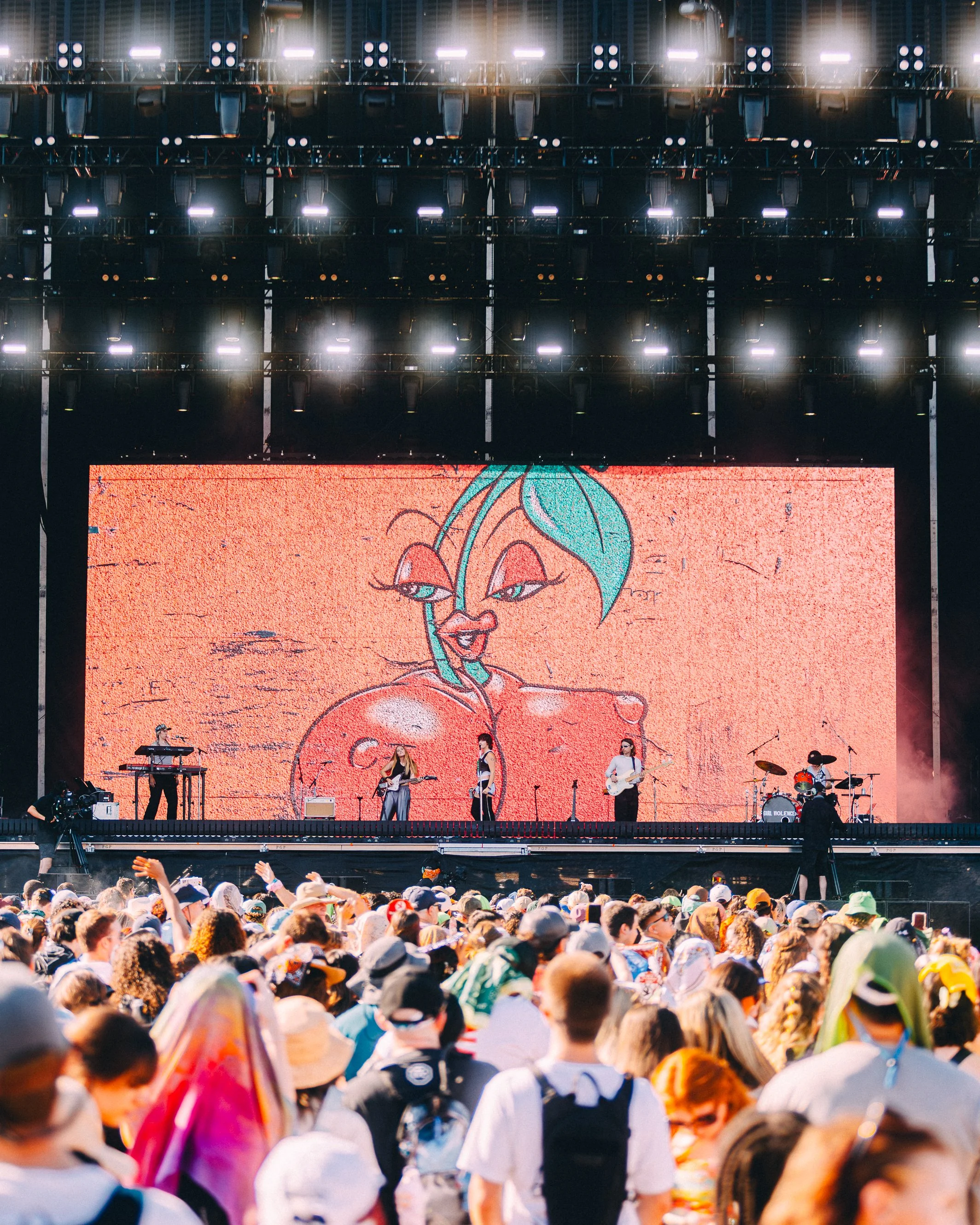 Concert stage with a digital screen displaying a cartoon character with teal hair and red lips, and a crowd of people watching and taking photos in the foreground.
