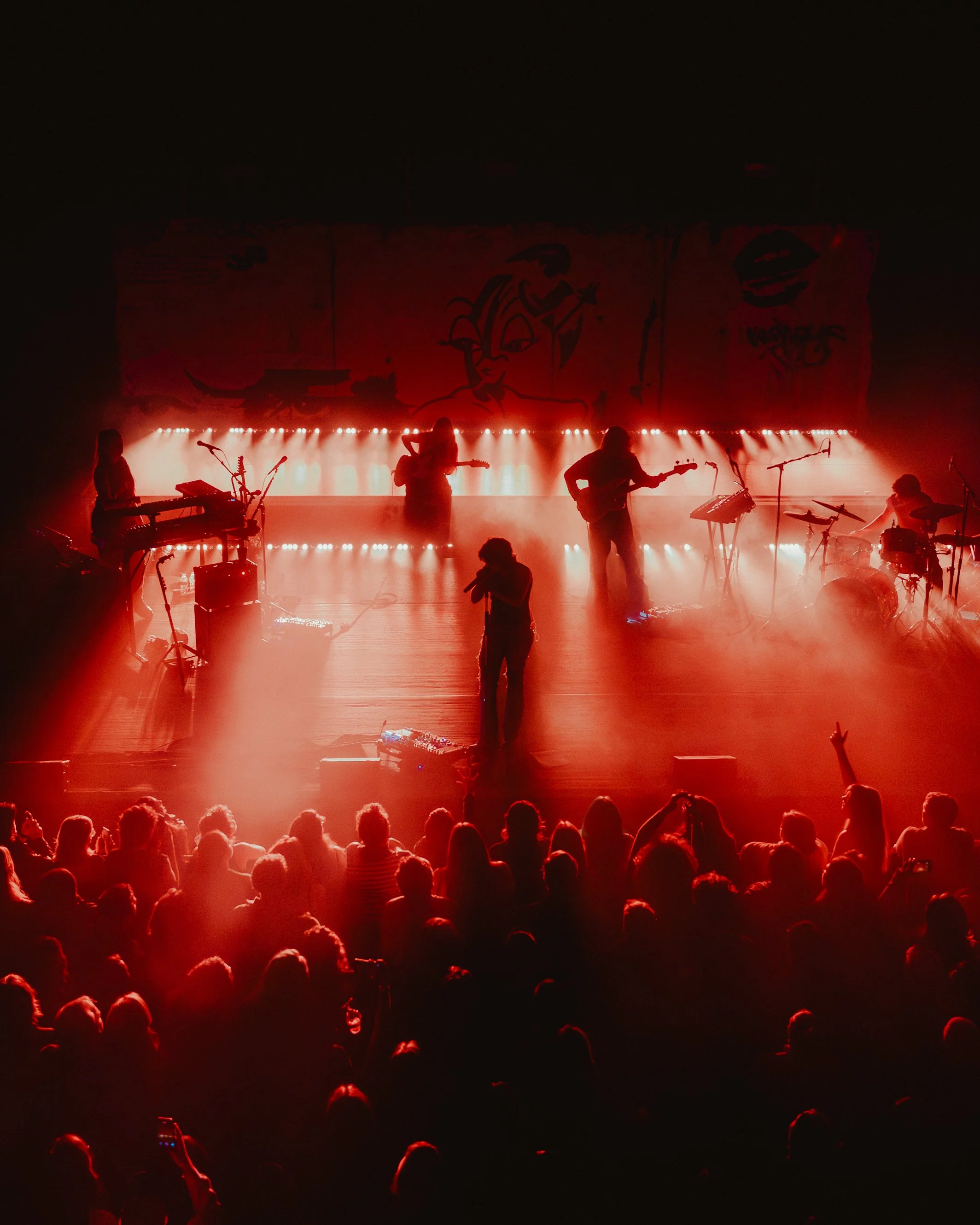 A concert stage illuminated with red lights, featuring multiple musicians playing instruments, with a large backdrop displaying abstract artwork.