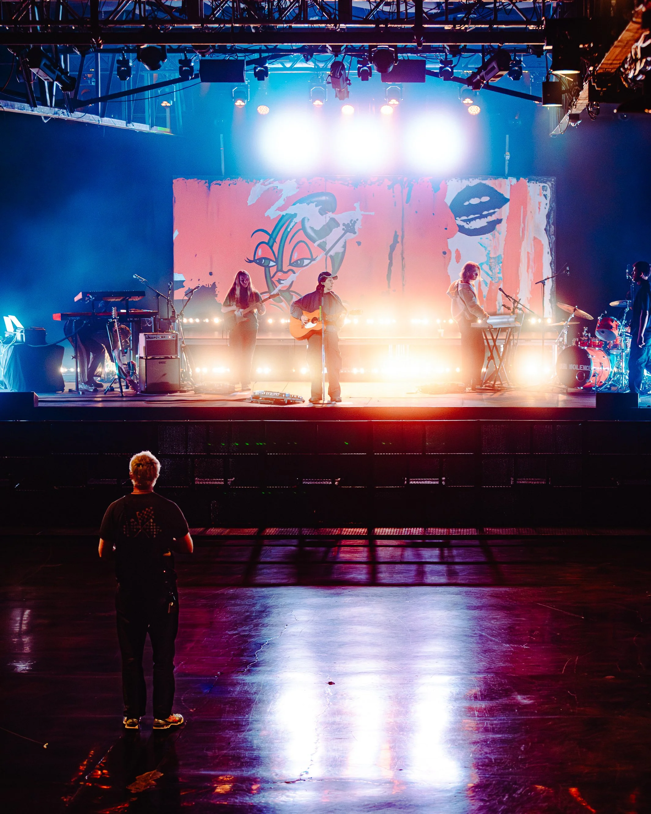 A group of musicians performing on stage with bright lights and a colorful backdrop, viewed from the audience area, with a person watching from the front.