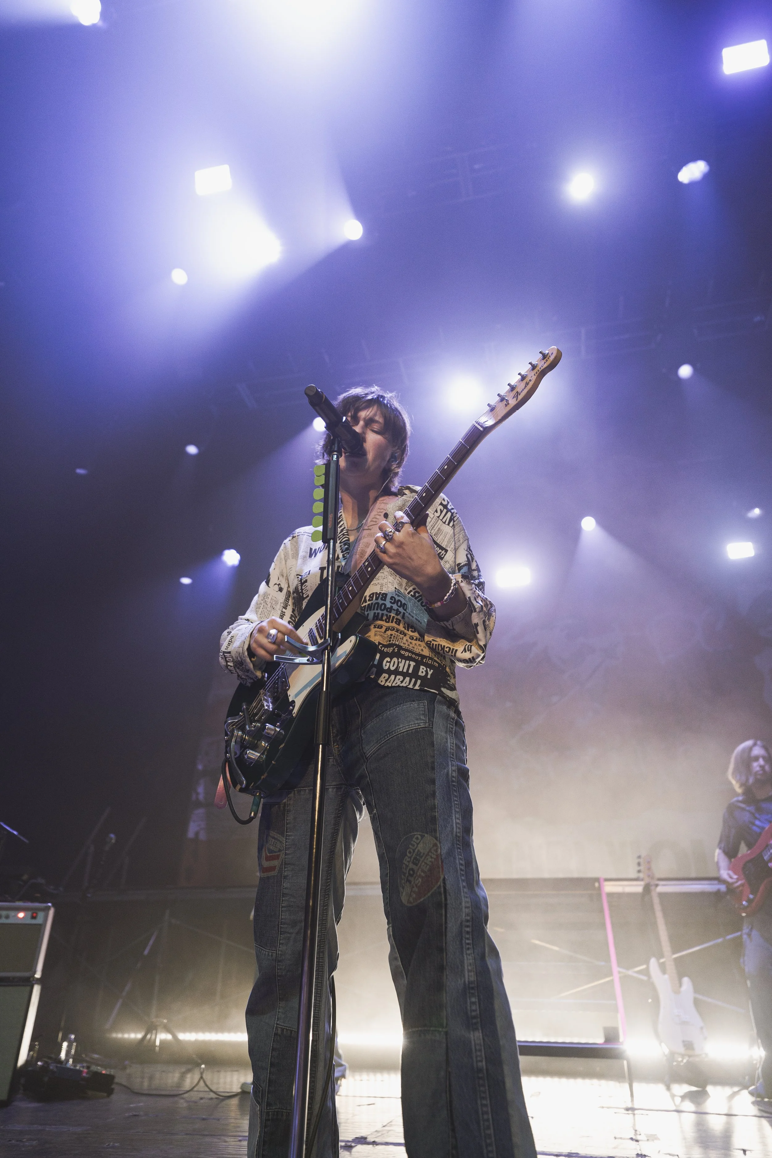 A musician performing on stage, singing into a microphone and playing an electric guitar, with stage lights shining bright overhead.