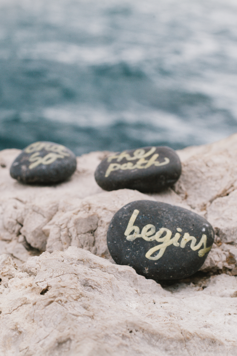 Rocks by the ocean with writing on them that say "and so the path begins".