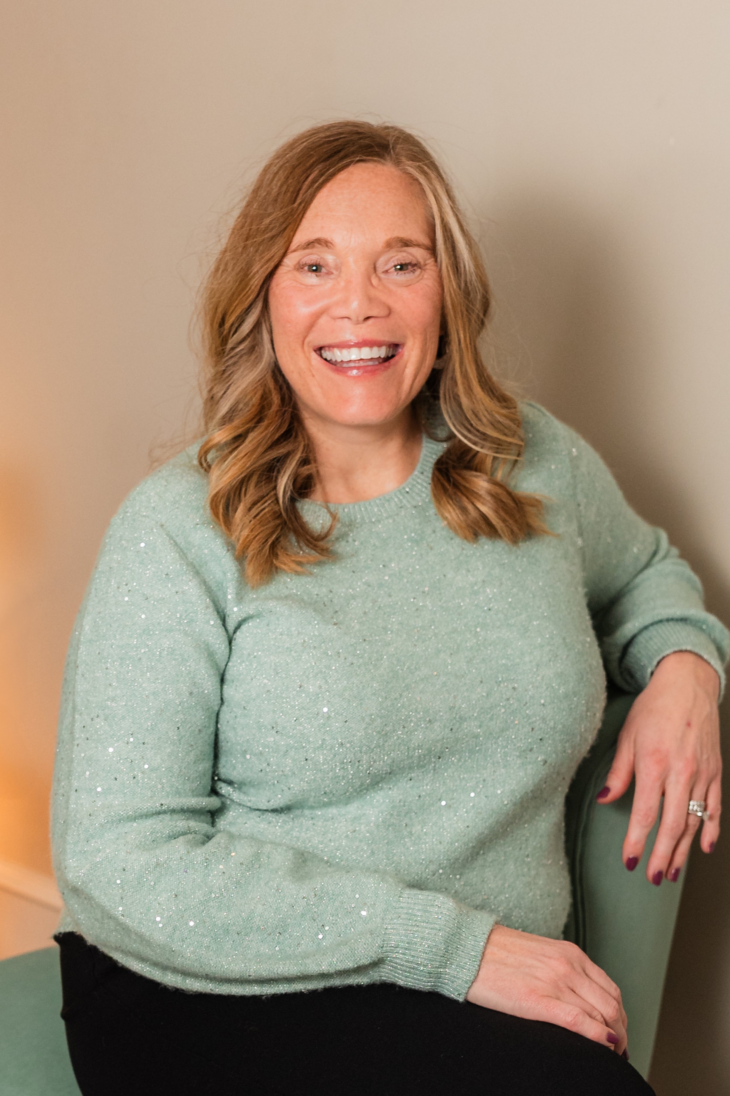 A woman with light skin, wavy red hair, and a big smile, sitting on a green chair against a neutral wall, wearing a light green sparkly sweater.