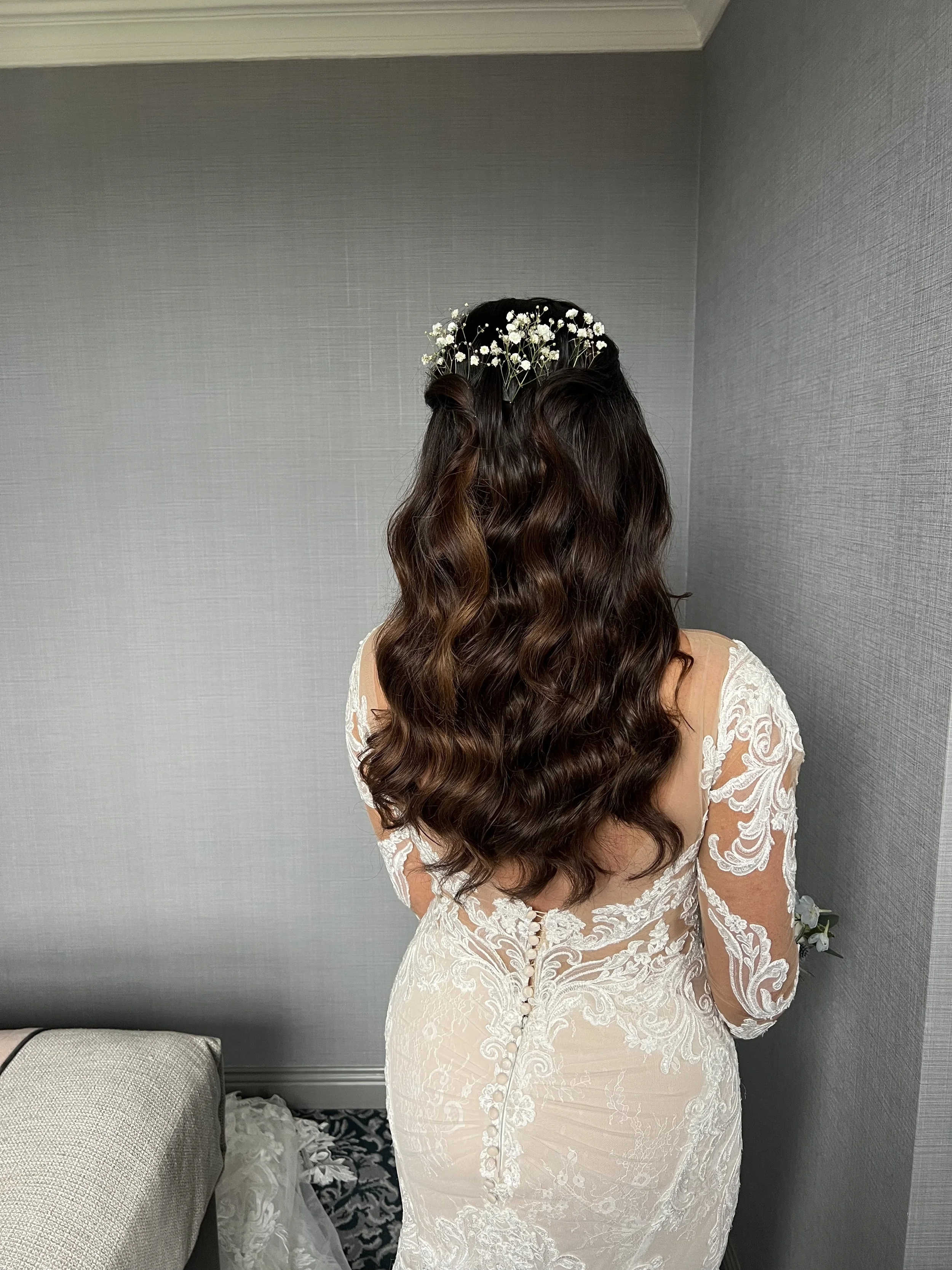 Four women dressed in formal attire, with three women in black gowns holding bouquets and one woman in a white gown, standing outdoors on a paved area with trees and a fence in the background, possibly at a wedding or special event.