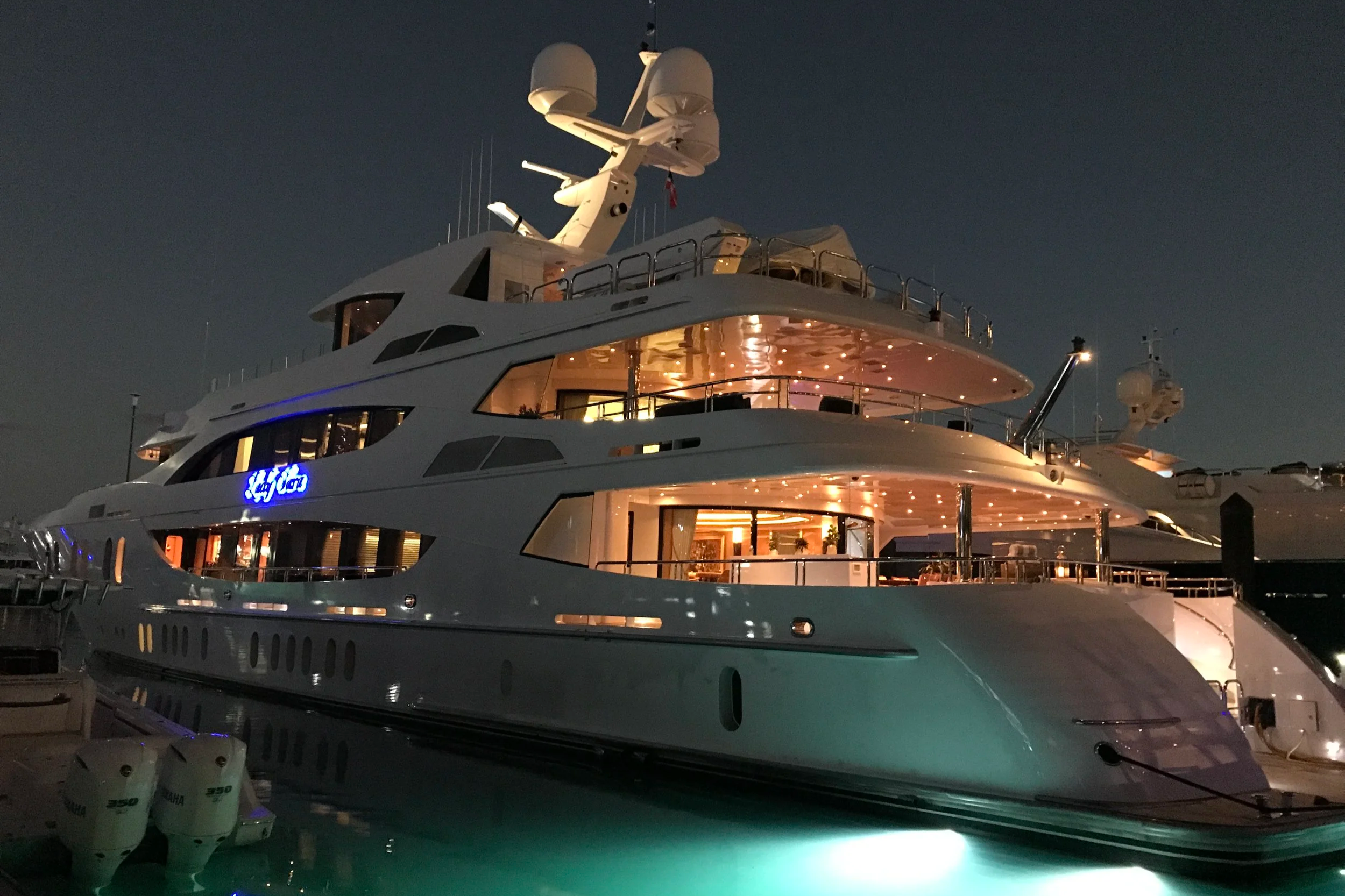 A large yacht docked at night with multiple decks illuminated from inside, showing windows and outdoor lighting. The sky is dark and the yacht's radar and communication equipment are visible on the top, with a partially visible boat in the foreground.