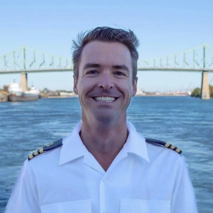 Smiling man in a white uniform with shoulder epaulets, standing in front of a river with a bridge in the background. Montreal, Yacht, St Lawrence,St-Laurent, Superyacht, agency, yacht agent