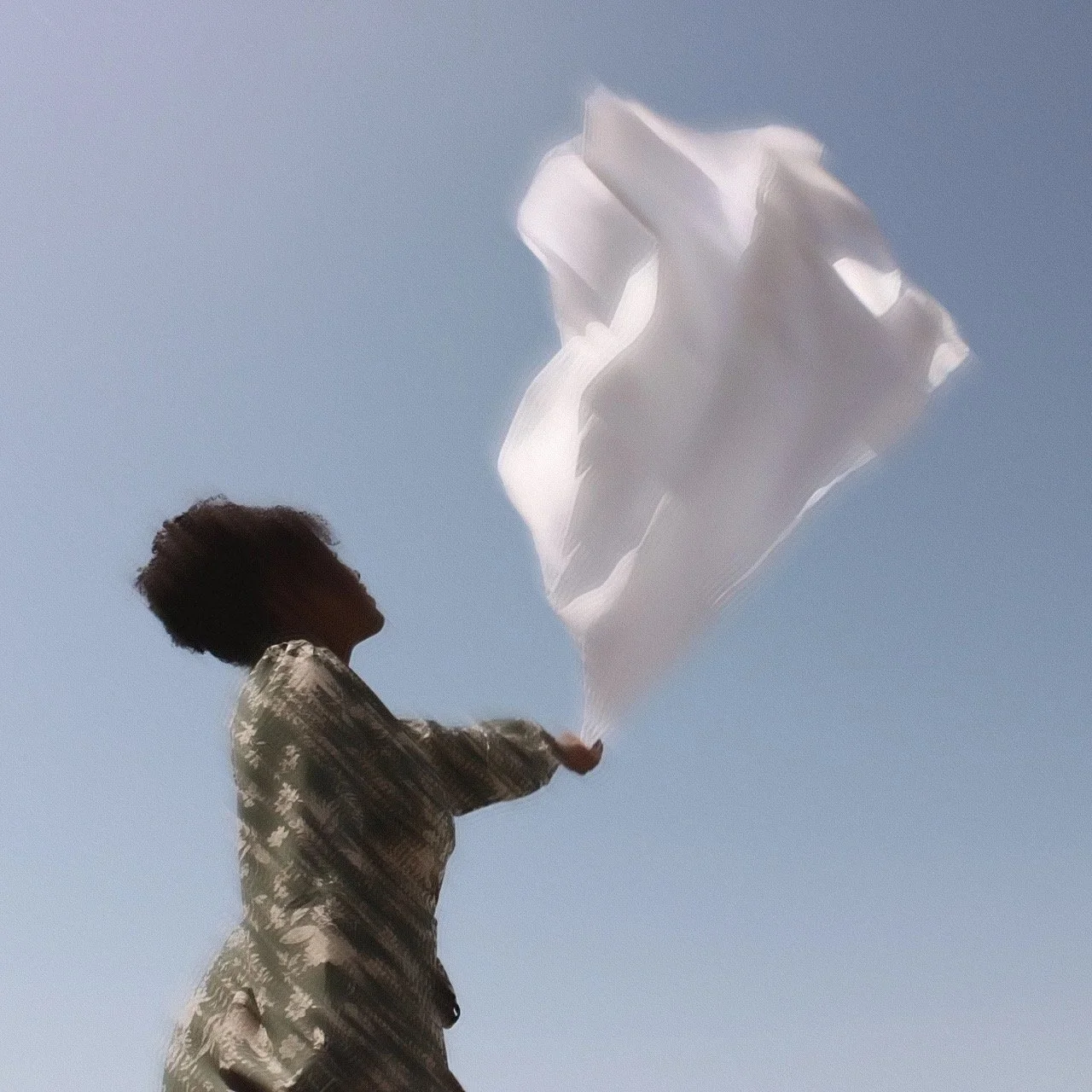 A woman with curly hair standing outdoors during the daytime, holding and letting a piece of white fabric or tissue paper blow in the wind against a clear blue sky.