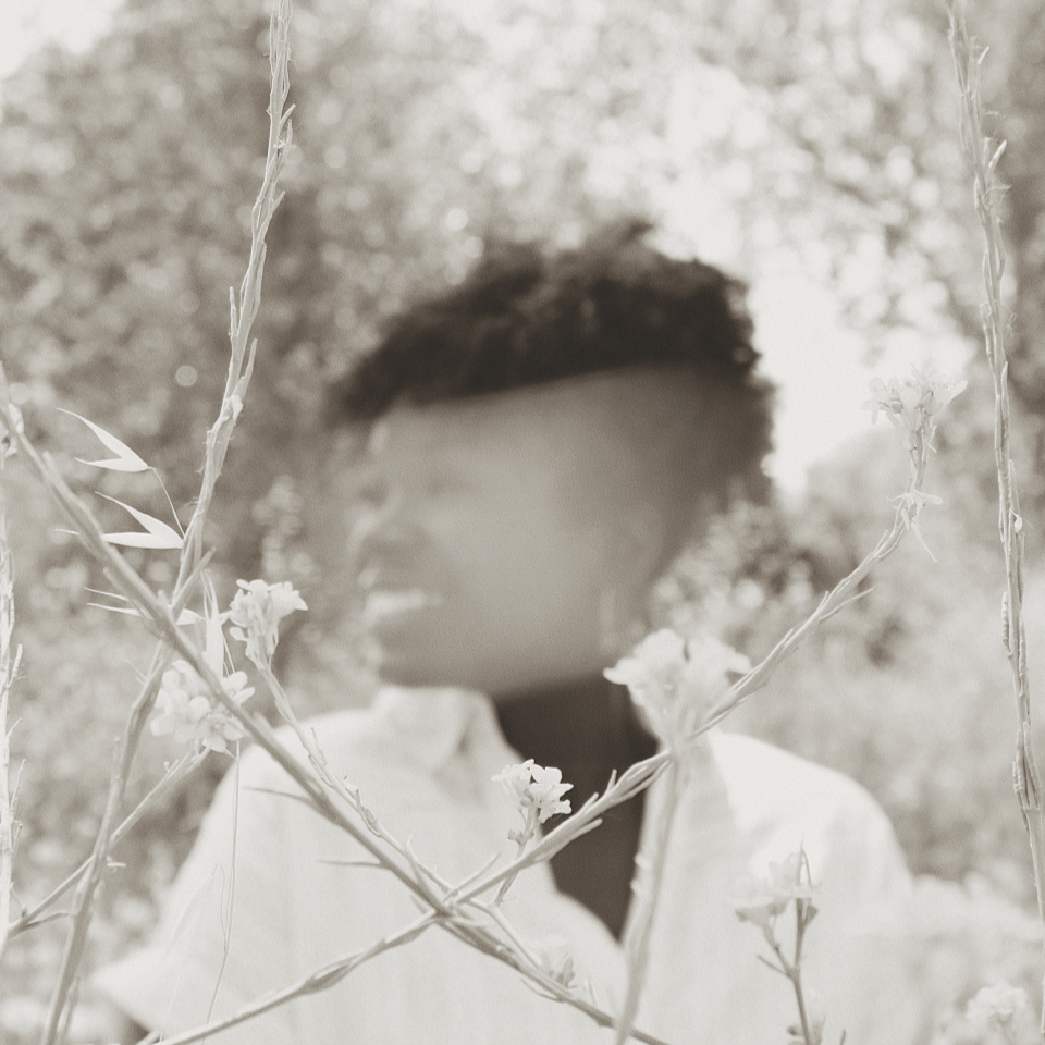 A person with short curly hair standing in a field of flowers, with trees in the background. The photo is in black and white.