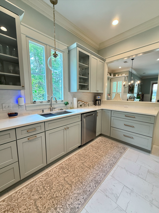 Modern kitchen with gray cabinets, white countertops, and stainless steel appliances. Features a large window, hanging light fixture, and a decorative patterned rug on the floor. Natural light illuminates the space in Beaufort, SC