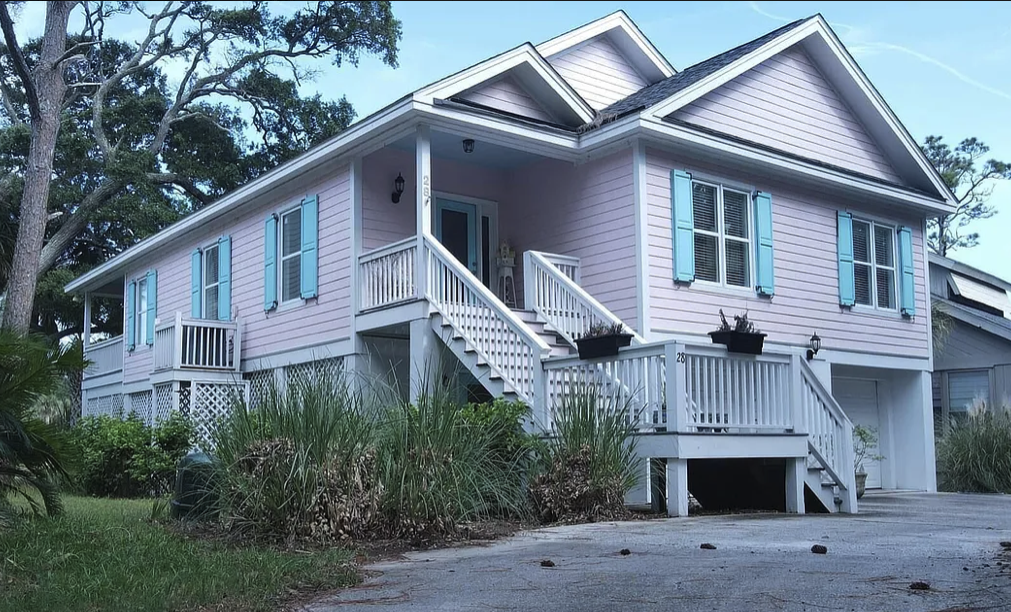 A raised light pink house with blue shutters and a white staircase, surrounded by trees and greenery.