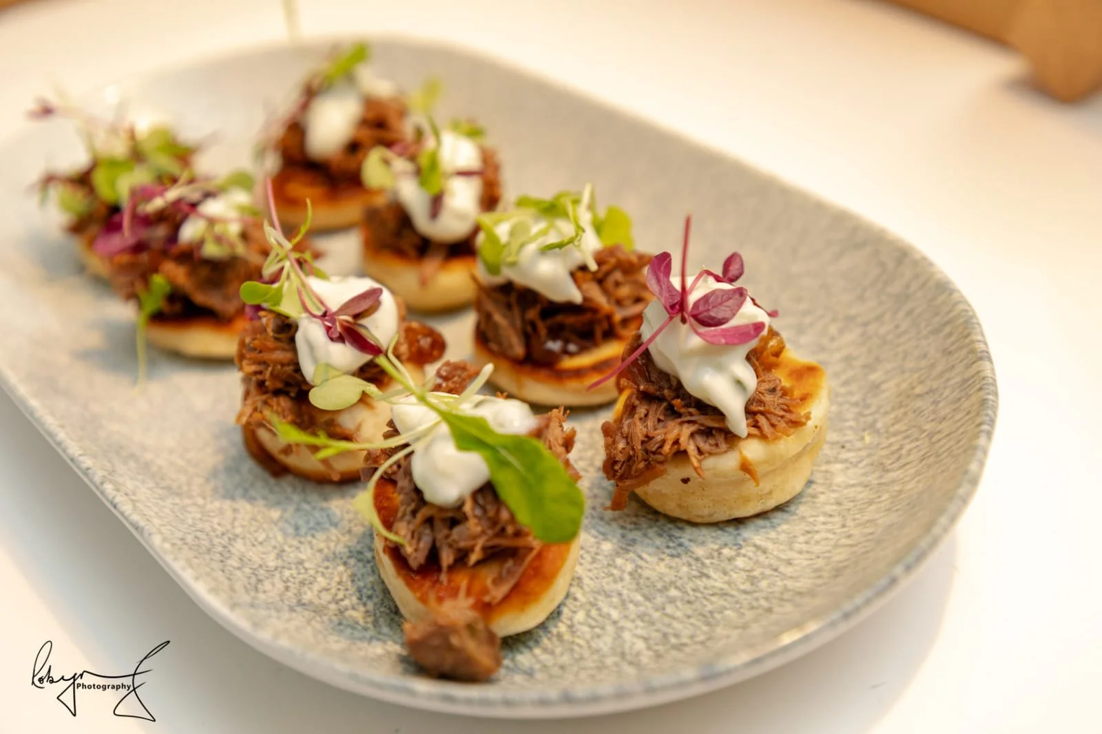 Bite-sized appetizers with shredded meat, dollops of white sauce, and microgreens on small toasted bread rounds, arranged on a decorative ceramic plate.