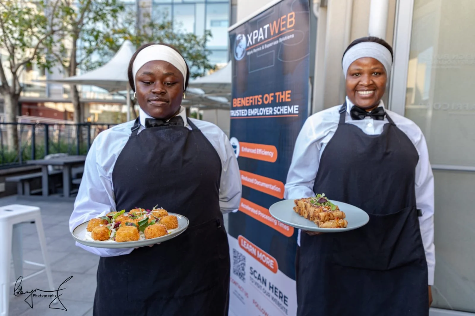 Two women in white shirts, black aprons, and headbands holding plates of food at an outdoor event, with a banner behind them displaying information about a web hosting scheme.