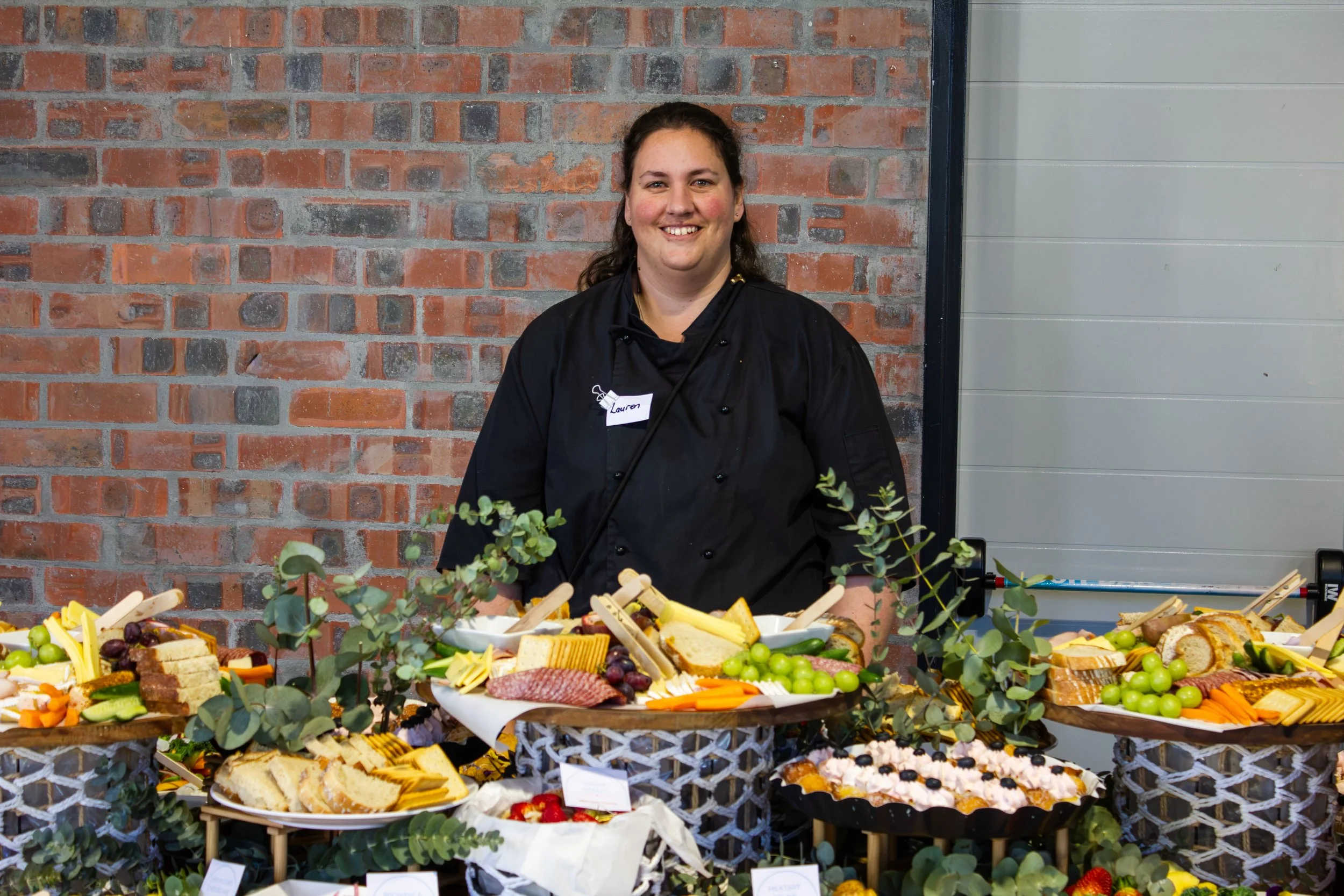 A woman dressed in a black chef's coat and apron, standing behind a table with an assortment of foods including meats, cheeses, vegetables, and grapes, with a brick wall in the background.