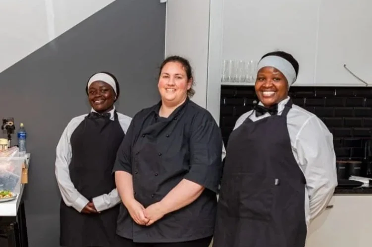 Three women, two in black aprons and chef hats, and one in a black chef coat, standing in a kitchen and smiling.