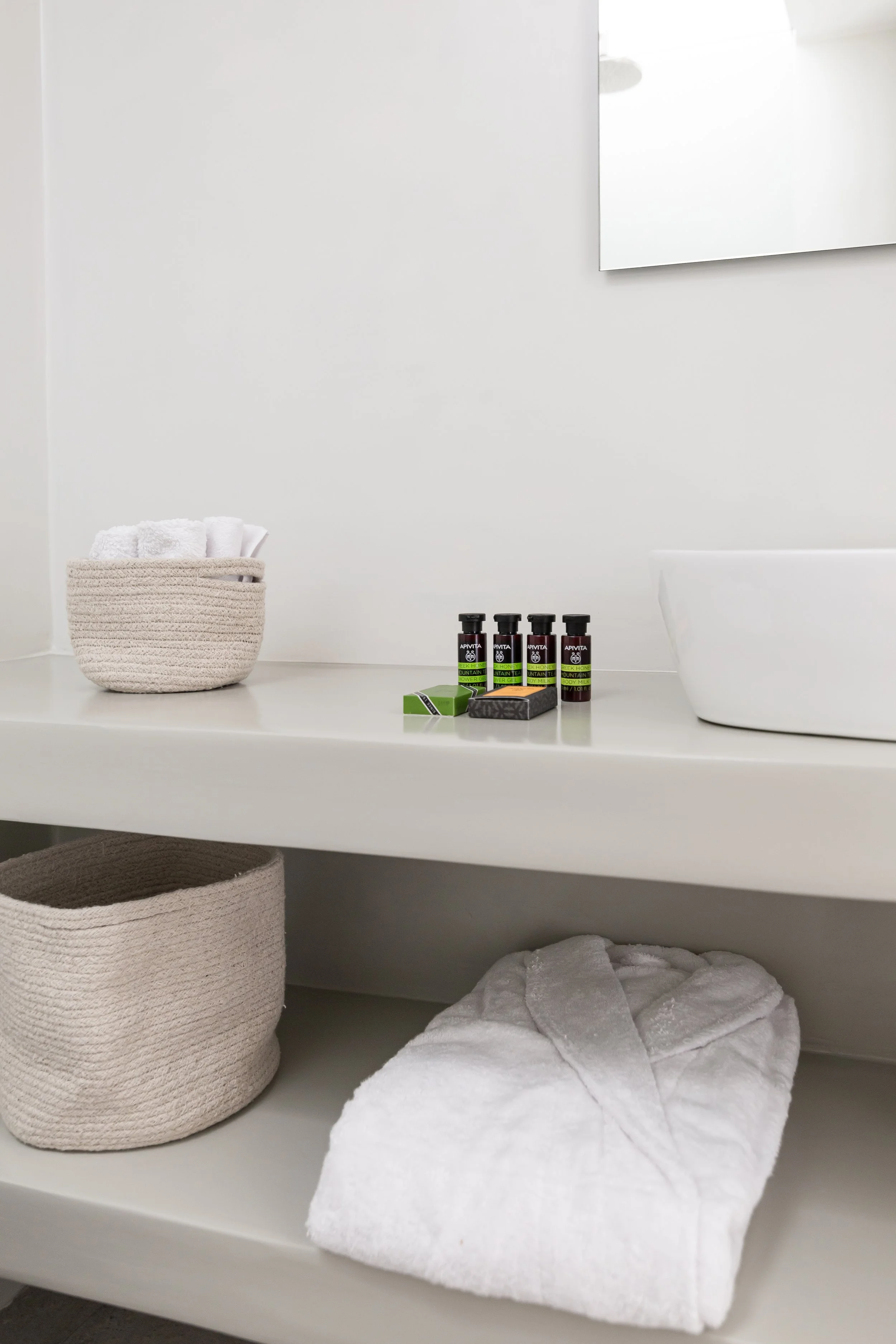 Bathroom shelf with a basket of rolled white towels, small bottles of toiletries, a box of soap, a white bathrobe, and a woven basket.
