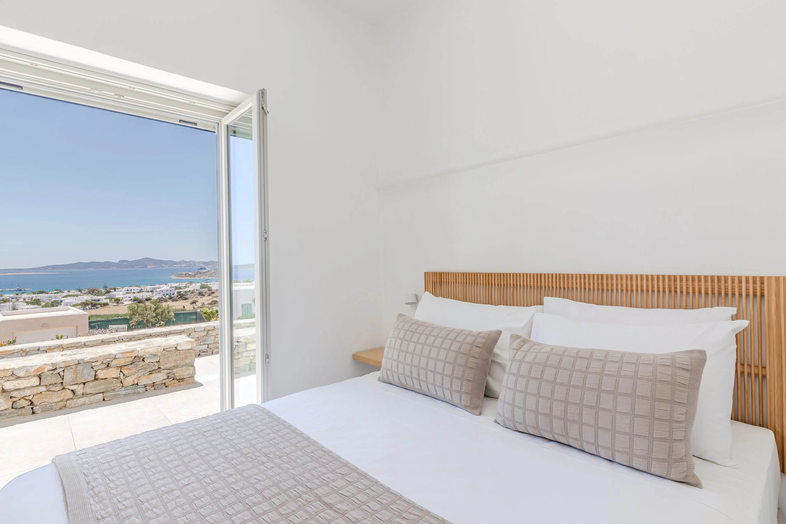 A bedroom with a bed that has white pillows and beige cushions, a wooden headboard, and an open door leading to a balcony with a view of the ocean and hills.