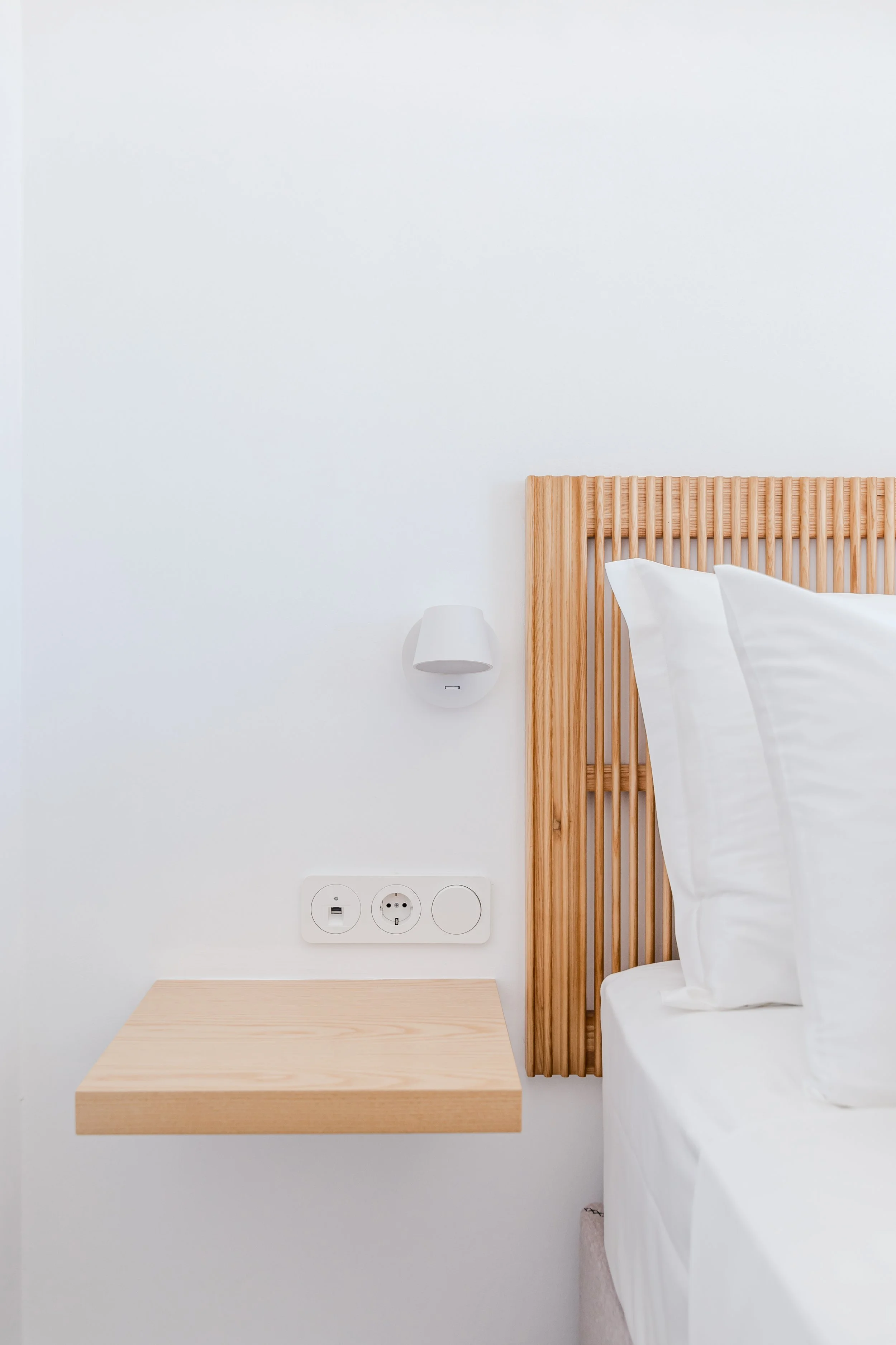A bedroom with a wooden headboard, white bedsheets and pillows, a beige bedside table, a wall-mounted small white lamp, and electrical outlets on a white wall.
