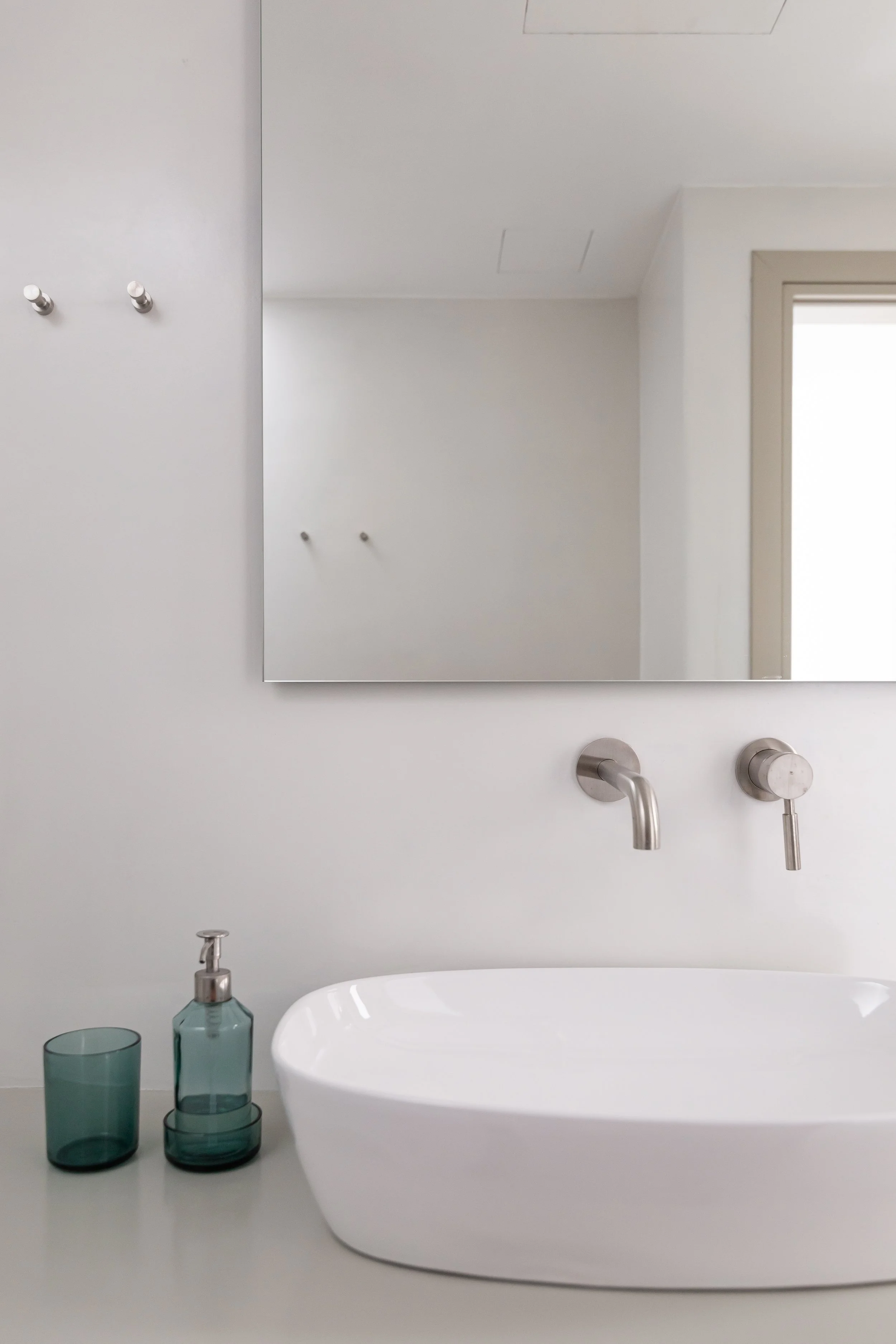 Minimalist bathroom with a white countertop, a white oval vessel sink, a wall-mounted faucet, a mirror, and two glass containers, one with a soap dispenser, against white walls.