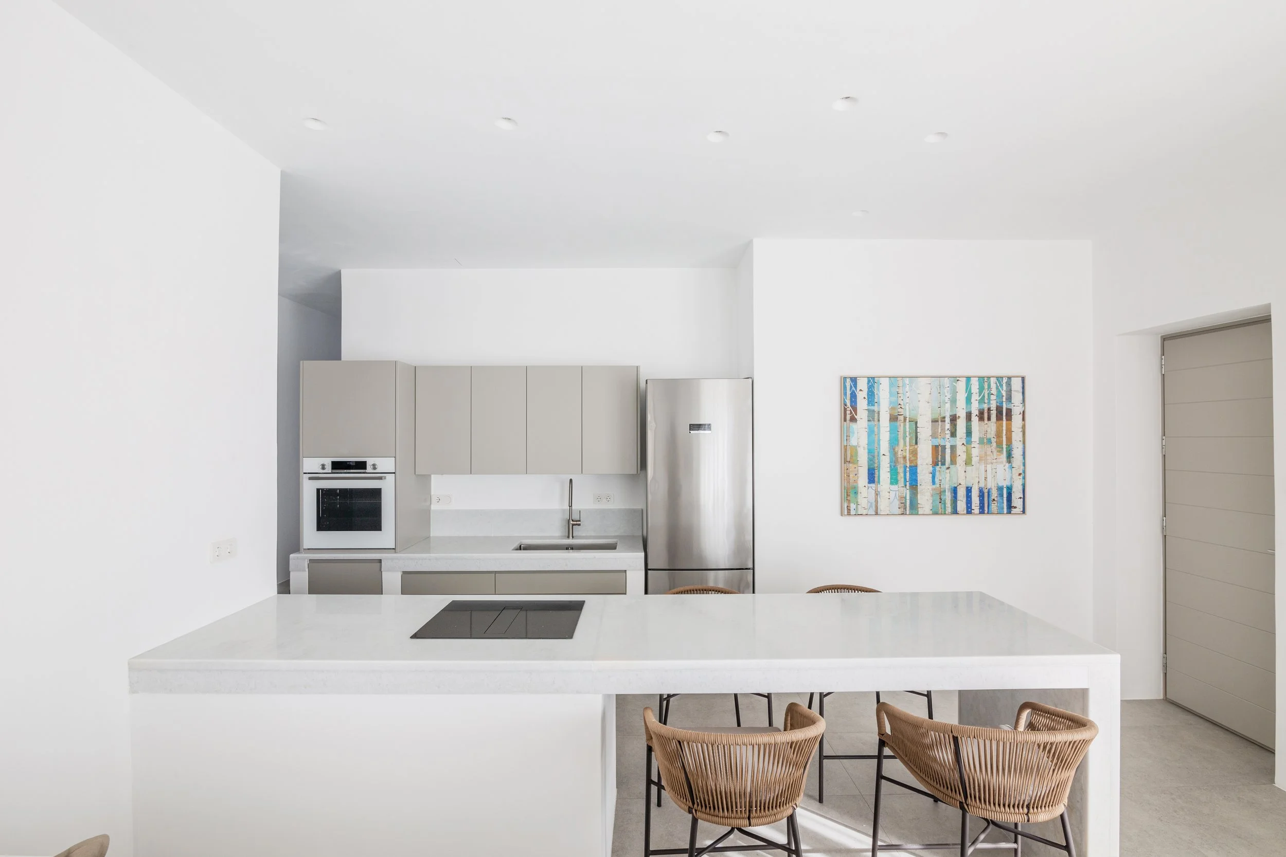 Modern minimalist kitchen with white walls, beige cabinetry, stainless steel refrigerator, built-in oven, gray countertop with sink, and a large white island with four woven chairs. A colorful abstract painting hangs on the wall near a gray door.