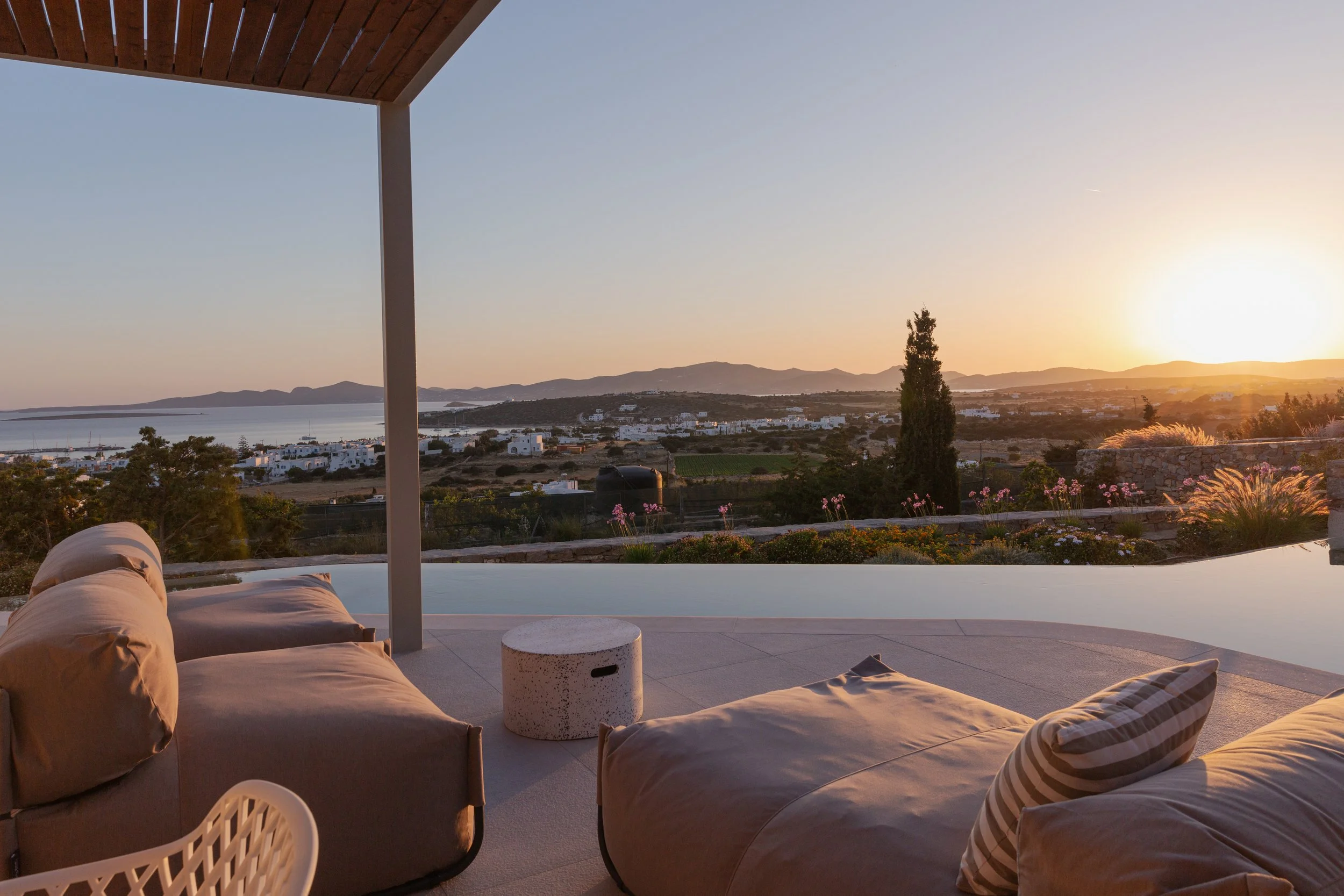 View from a terrace or porch at sunset, overlooking a landscape with water, white buildings, trees, and mountains in the distance. There are cushioned chairs and a small table in the foreground.