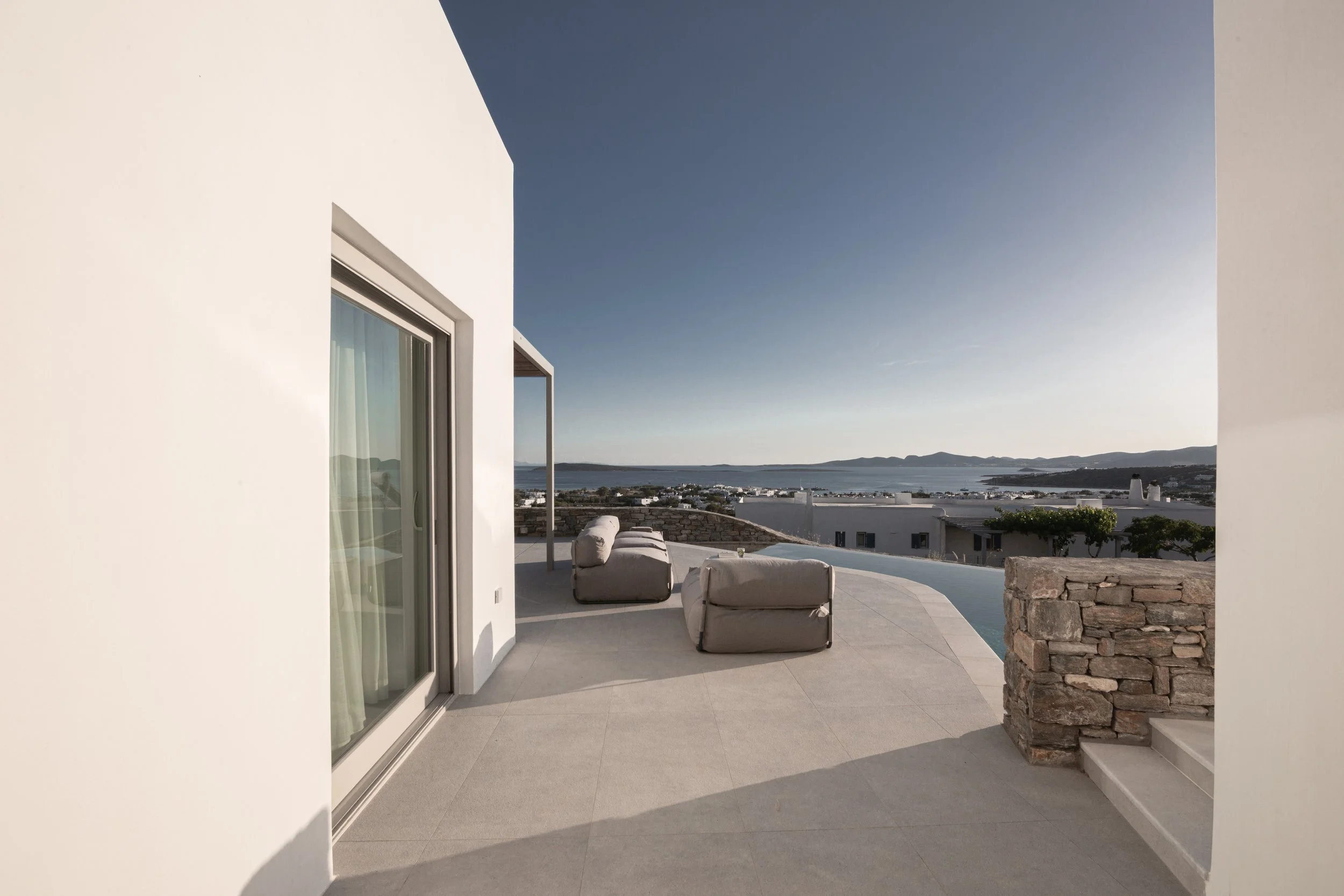 Modern white building with large sliding glass door leading to outdoor patio with lounge chairs, overlooking a body of water and distant hills under a clear blue sky.