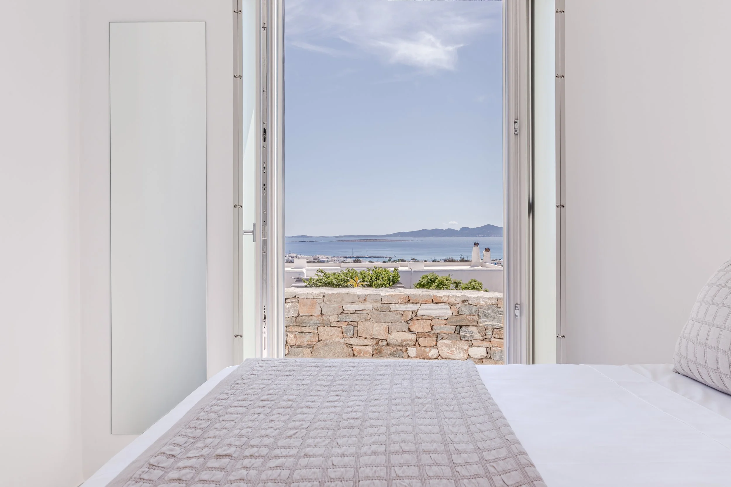 View from inside a white bedroom looking out of an open balcony door to a scenic seaside landscape with white buildings, greenery, blue water, and distant mountains under a partly cloudy sky.