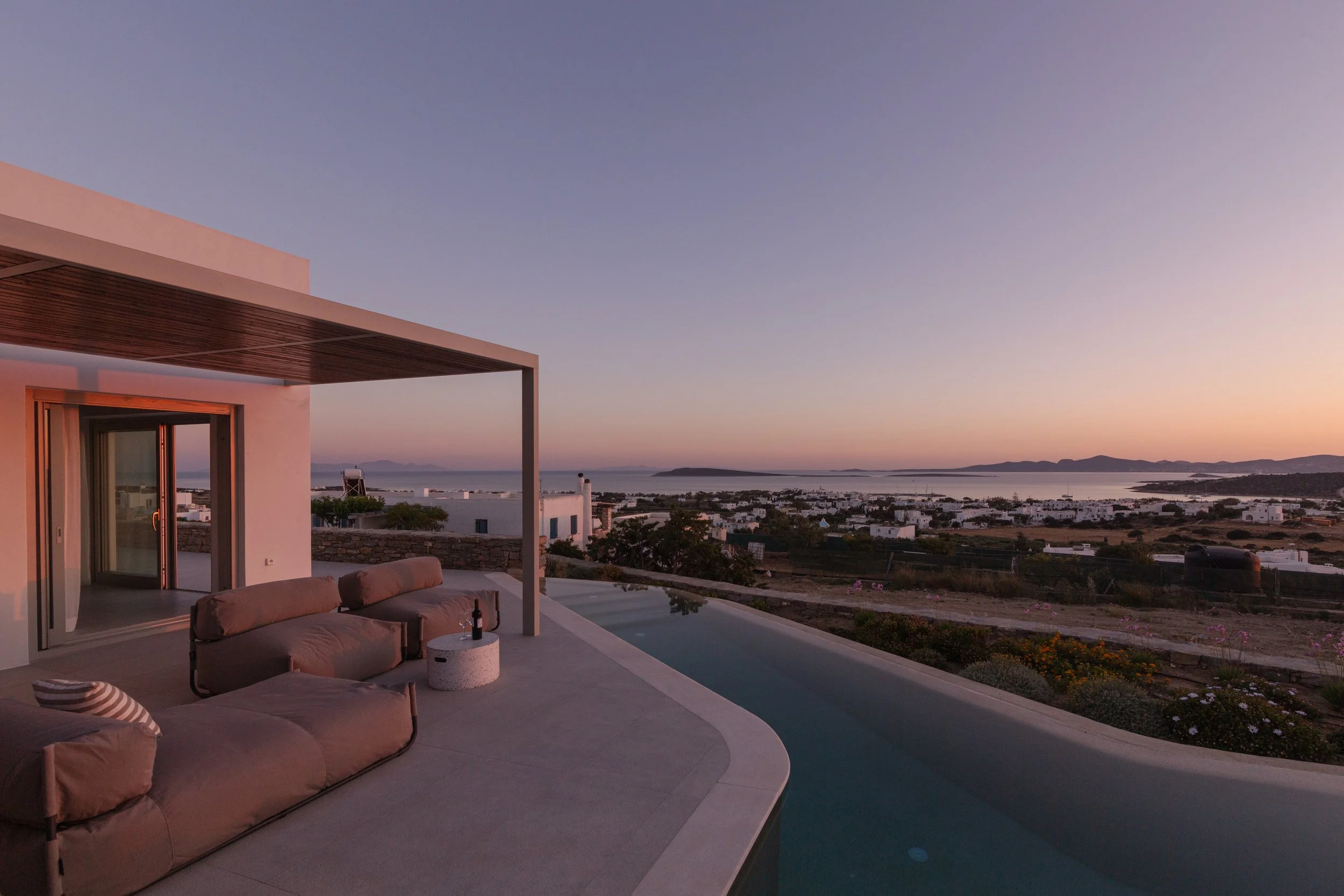 Modern outdoor patio with beige sofas, a wine bottle, and a small table, overlooking a sunset view of a coastal village with white buildings, water, hills, and sky.