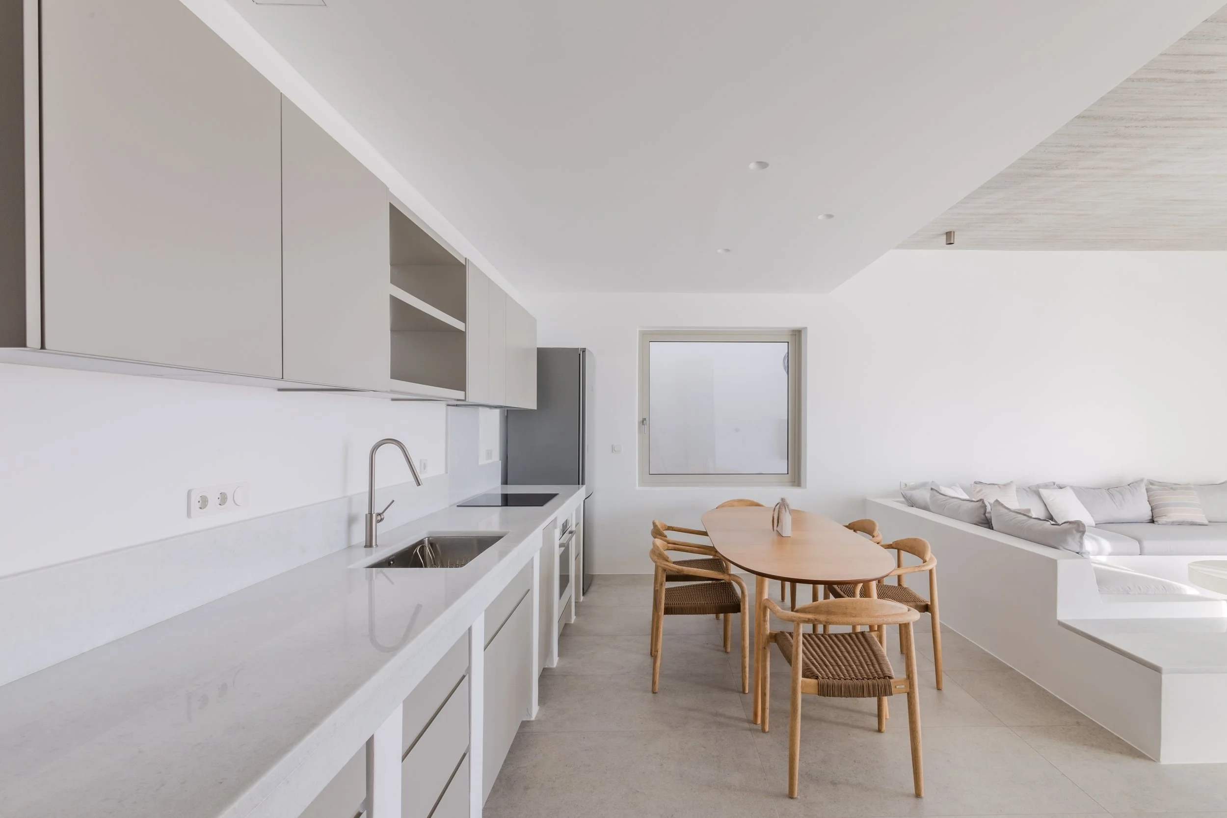Minimalist open-plan kitchen and dining area with white cabinetry, a wooden dining table with six chairs, and a white sectional sofa with cushions in a bright room