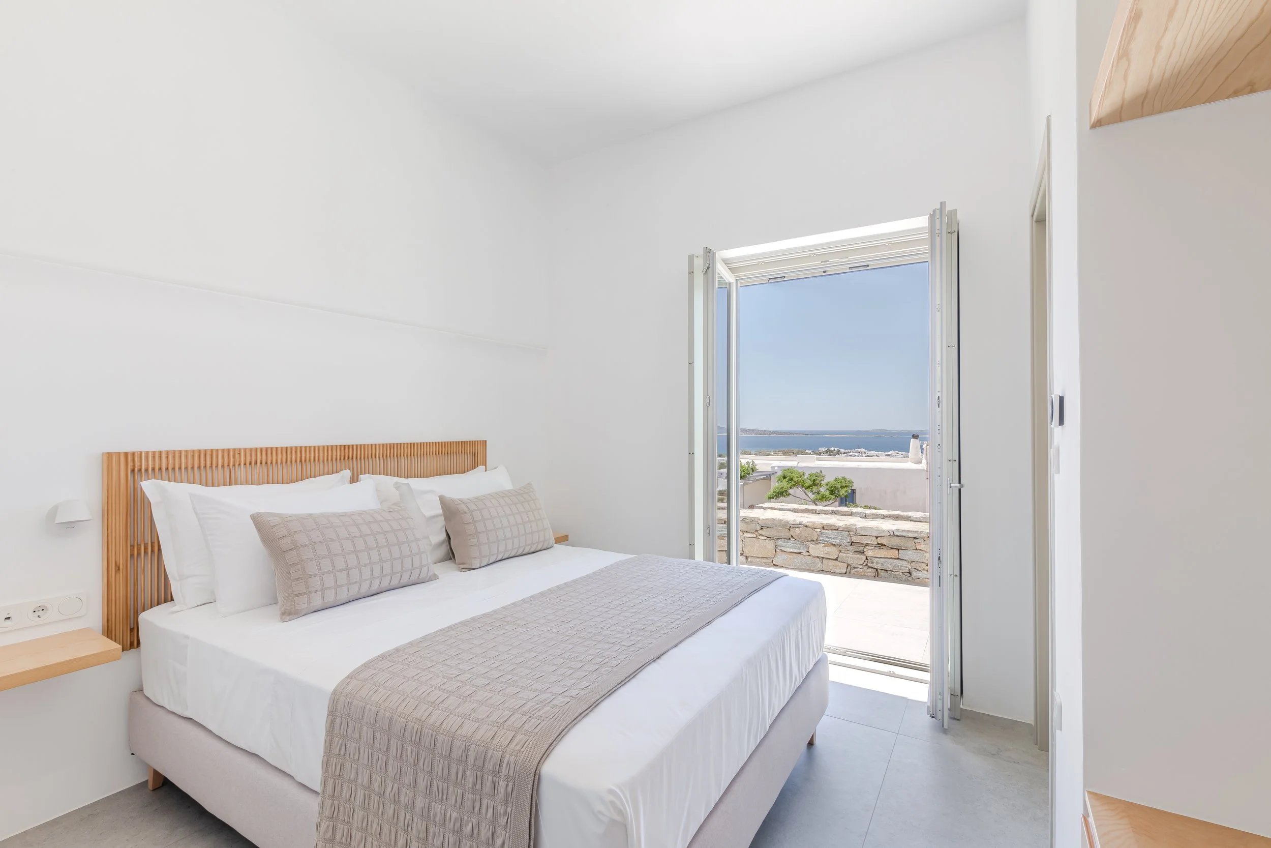 A bright, minimalistic bedroom with a large bed, beige pillows, and an open door leading to a balcony with a view of the ocean and clear blue sky.