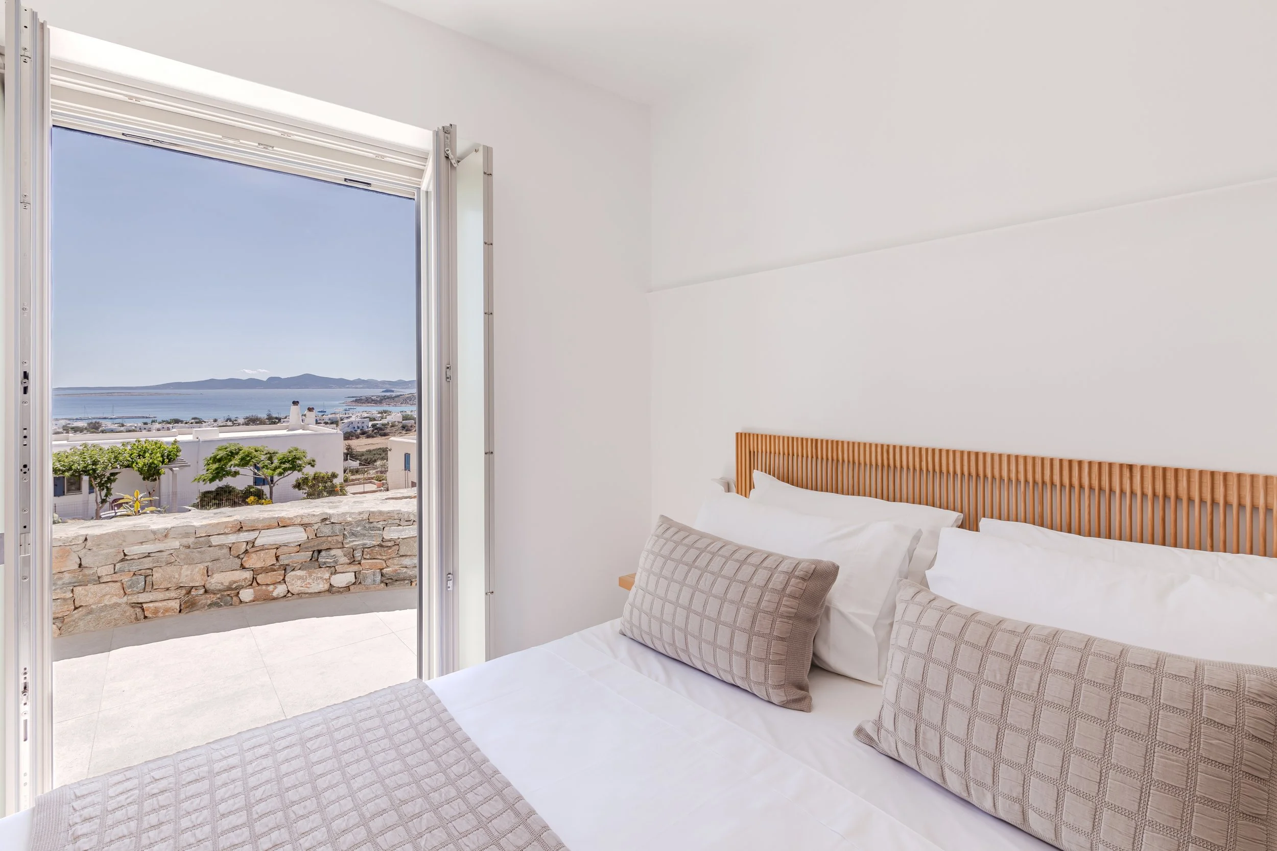 A bedroom with a bed having white linens and beige checkered pillows, next to an open door leading to a balcony with a view of a seaside town and mountains in the distance.