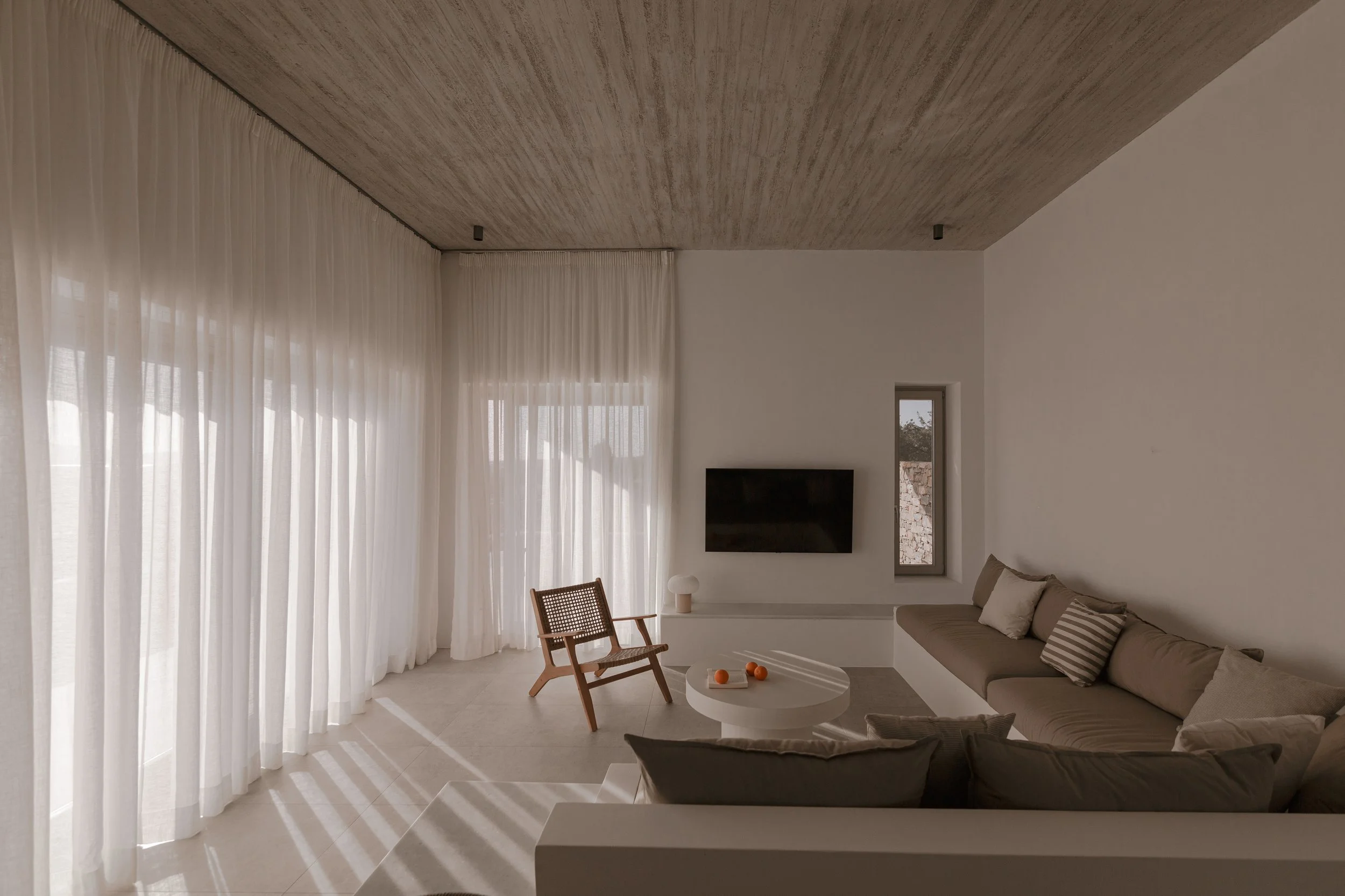 Minimalist living room with white sheer curtains, a beige sectional sofa, a wooden chair, a round white coffee table with oranges, a wall-mounted flat-screen TV, a small table lamp, and a narrow window showing an outdoor stone wall.