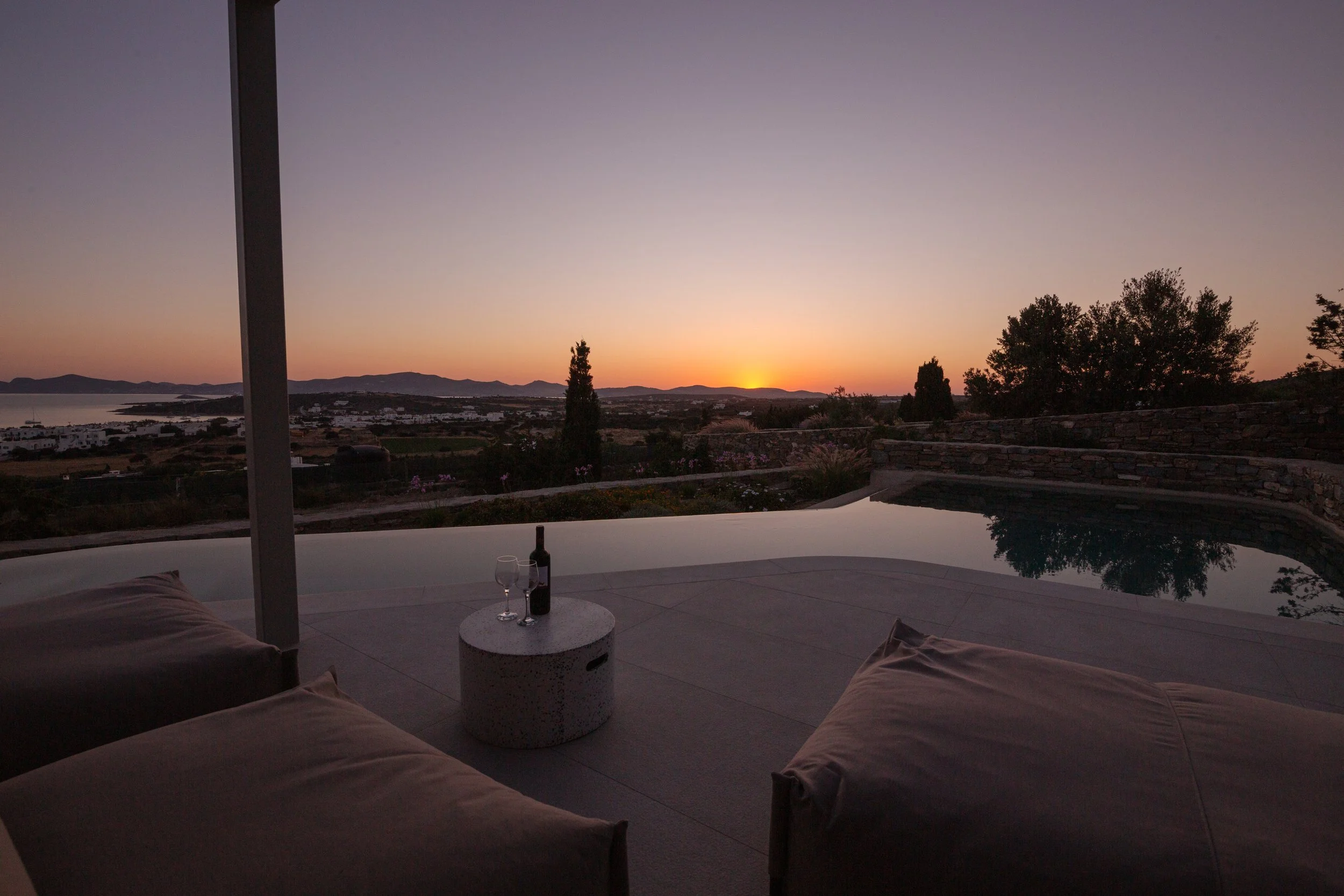 A sunset view from a patio with lounge chairs, a small round table with a bottle of wine and two glasses, an infinity pool, and a scenic landscape with hills, trees, and water in the background.