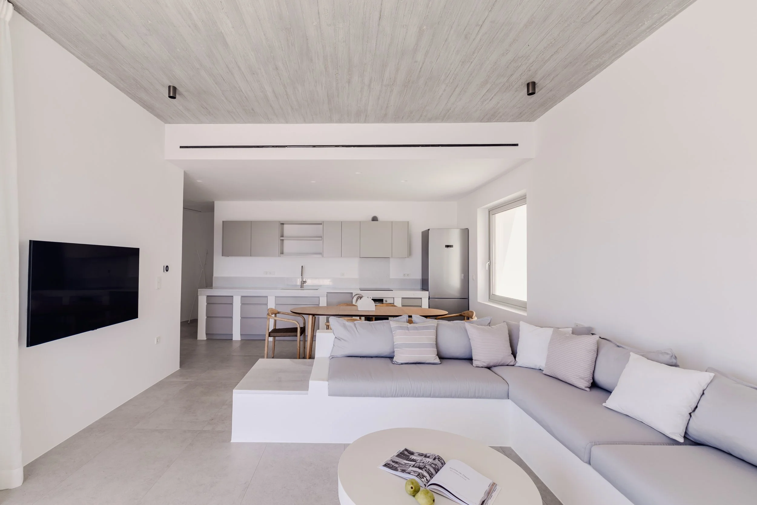 Modern living room with white walls, gray sectional sofa with pillows, a small round coffee table with an open book and two green apples, and a kitchen in the background with light gray cabinets, a white countertop, and a stainless steel refrigerator