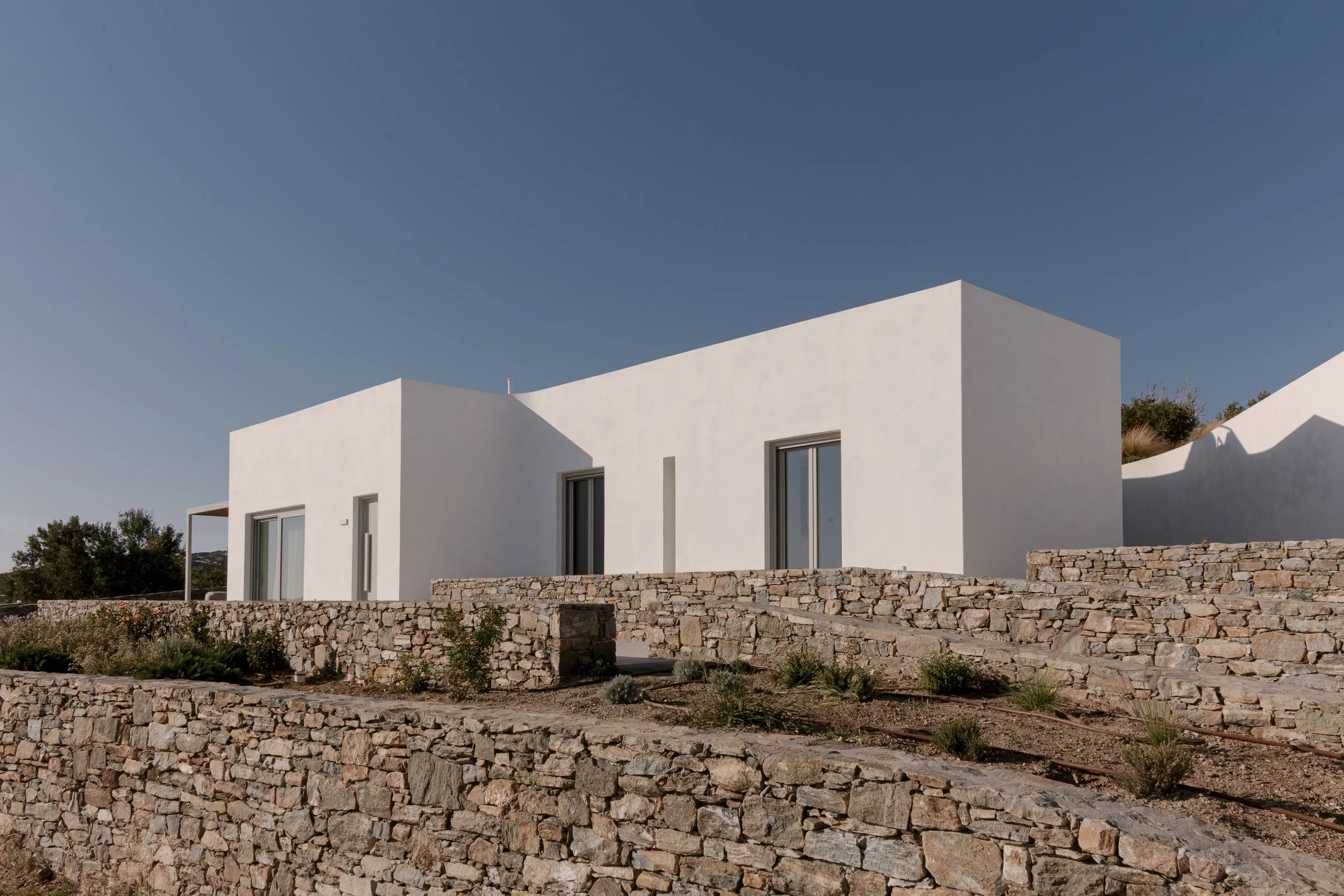 Modern white house with flat roof and large windows, built on a multi-level stone terrace with small plants, under a clear blue sky.