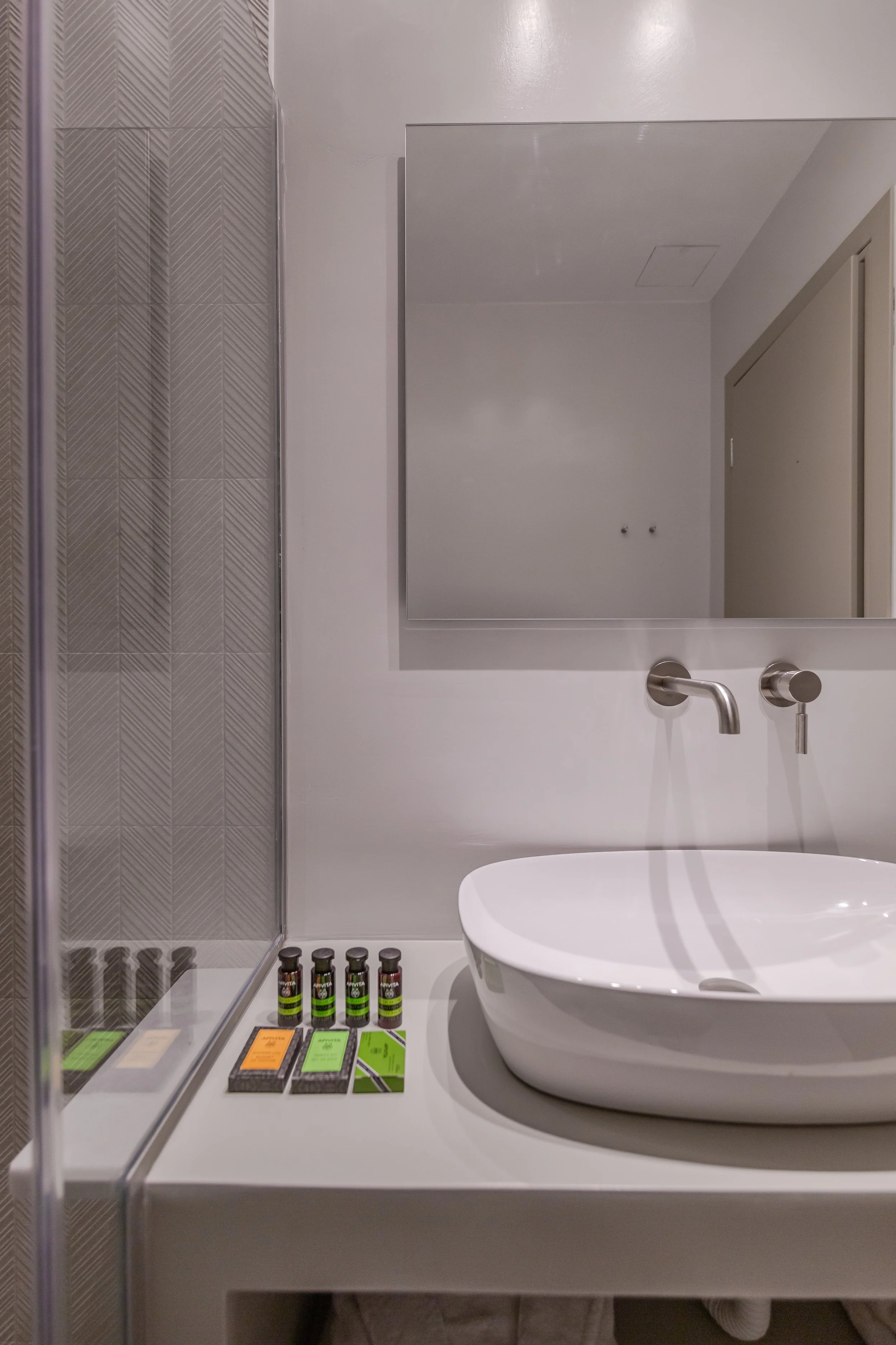 A modern bathroom sink area featuring a large mirror, a white oval vessel sink, a wall-mounted faucet, and toiletries placed on the counter, with tiled wall on the left.