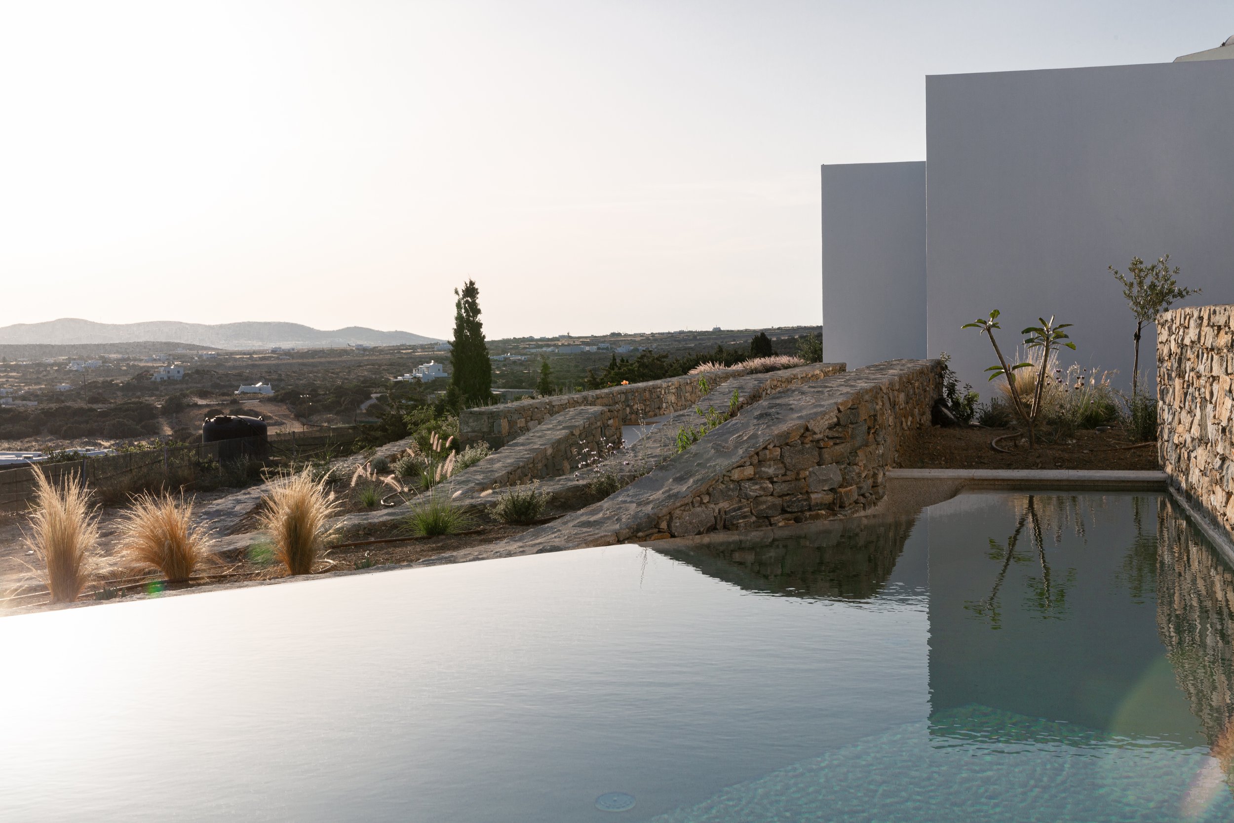 A modern outdoor swimming pool overlooks a hilly landscape with distant mountains. The pool is surrounded by a stone wall and desert plants, with a minimalist white building in the background. The sky is clear with bright sunlight.