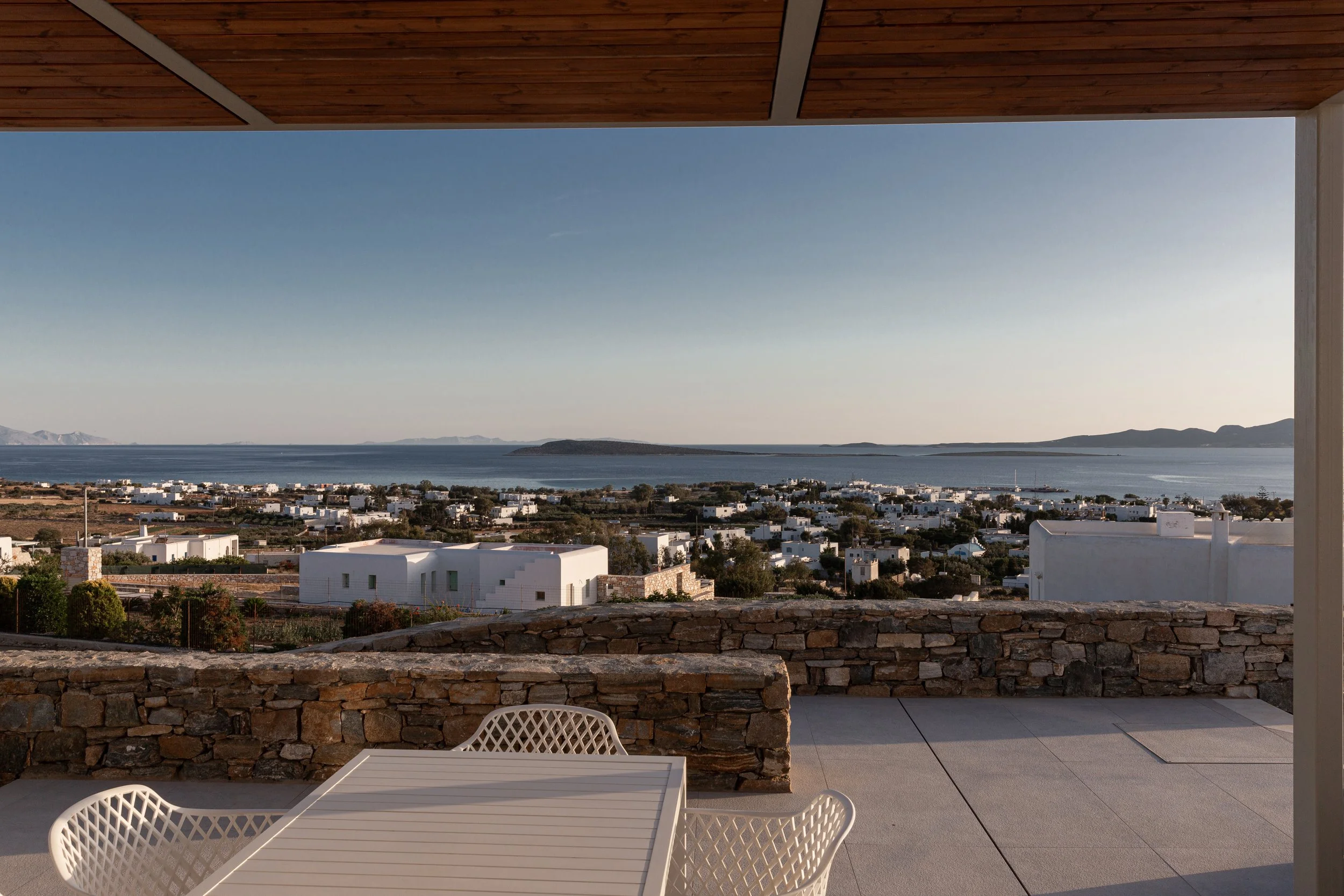 View of a coastal town with white buildings, water, and distant mountains, seen from a terrace with a stone wall and outdoor furniture.