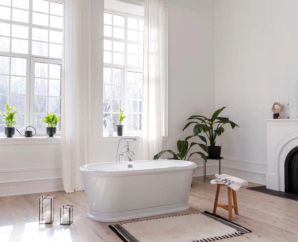Bright bathroom with a white freestanding bathtub, large windows with white curtains, potted plants on the windowsill, a large green plant near the fireplace, a small stool with towels, two black candle holders with candles, and a rug on light wood flooring.