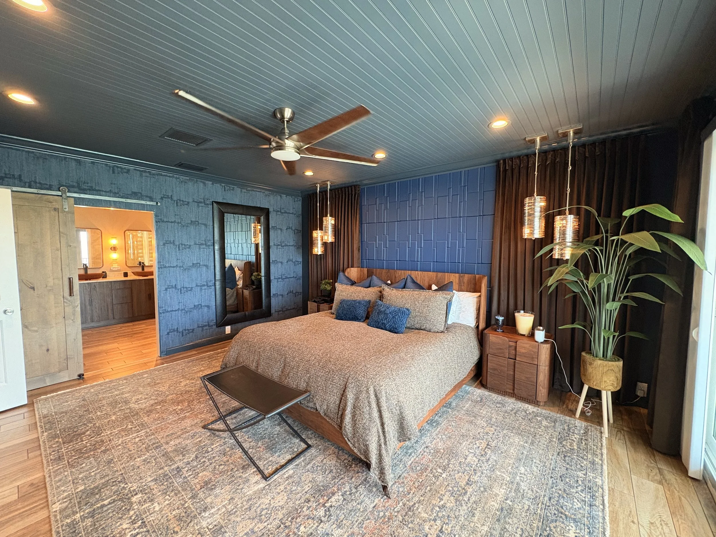 Completed primary bedroom design for Seabreeze Modern in Gulfport, Florida featuring moody blue wall paneling, walnut furnishings, layered lighting, textured textiles, and a warm modern retreat aesthetic.